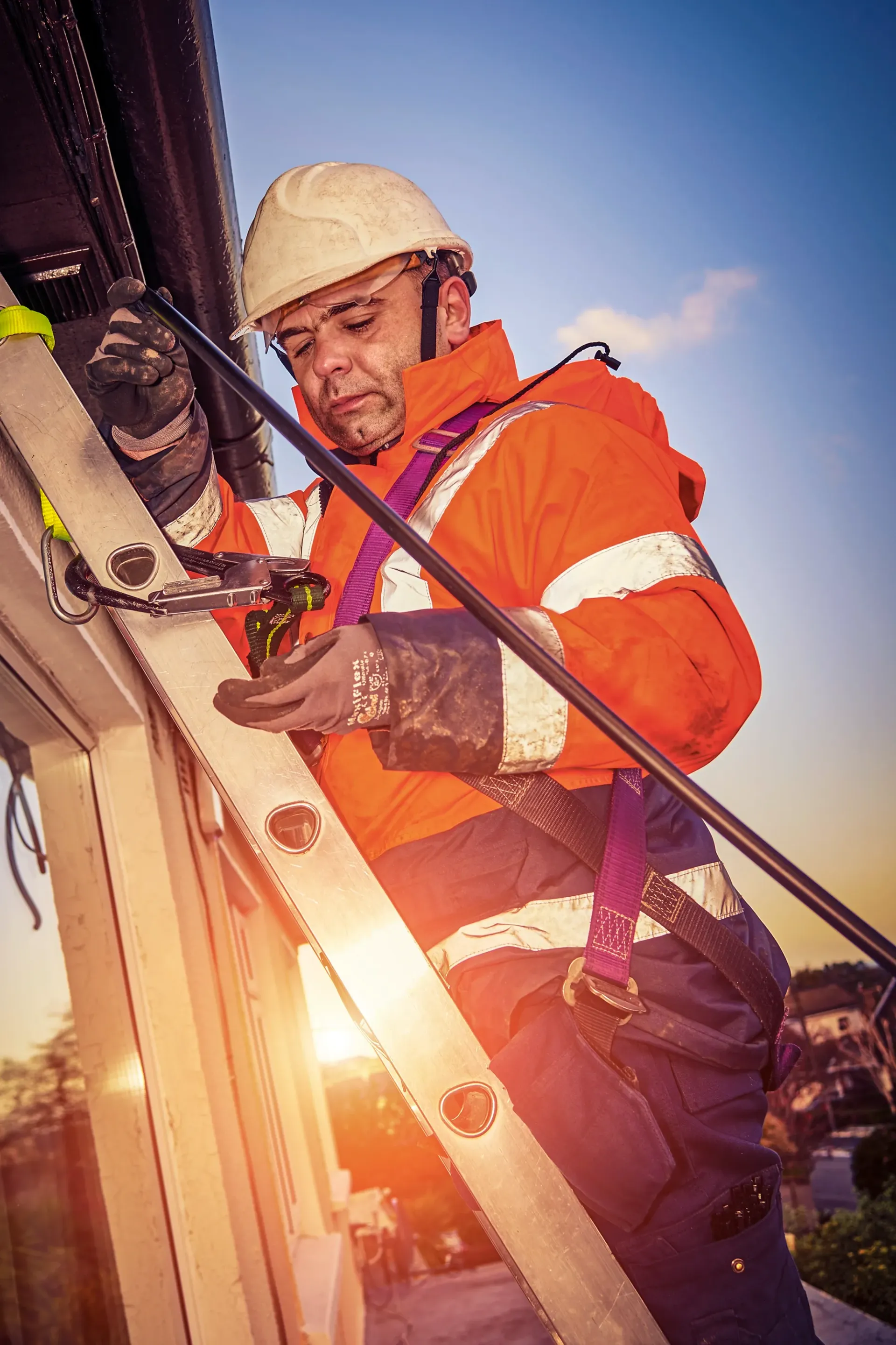 Construction worker in safety gear on a ladder installing something near the roofline; sunny, daytime.