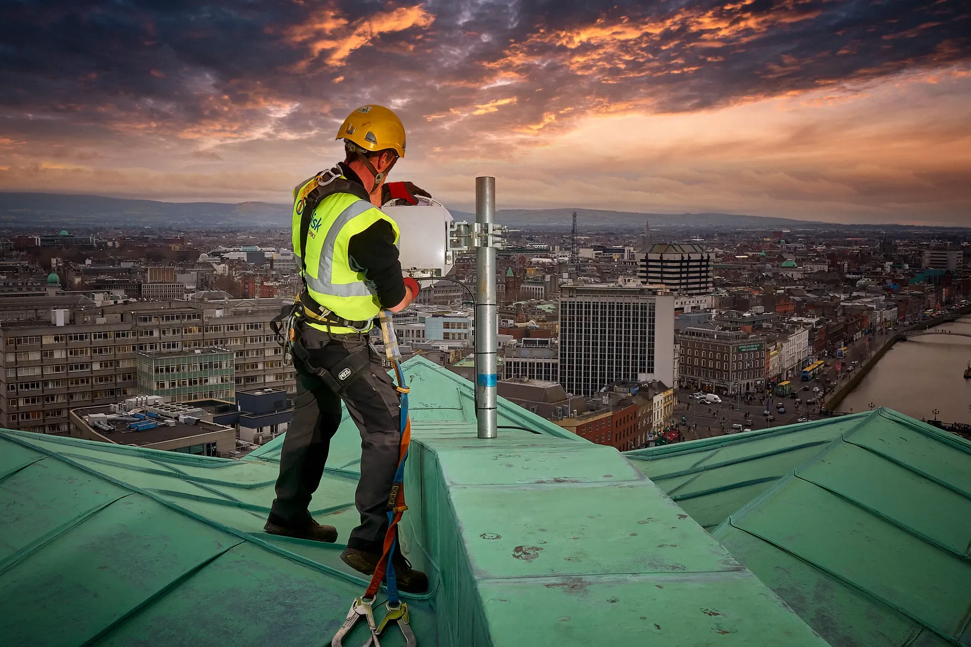 Person in safety gear installs equipment on a rooftop with city views and sunset sky.