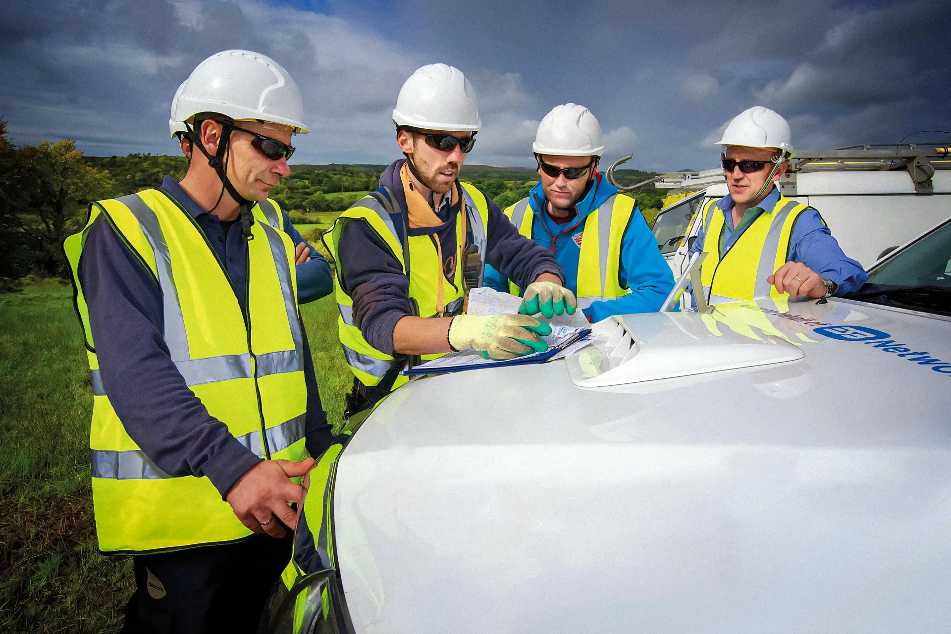 Four workers in safety vests and hard hats reviewing paperwork on a white vehicle outdoors.