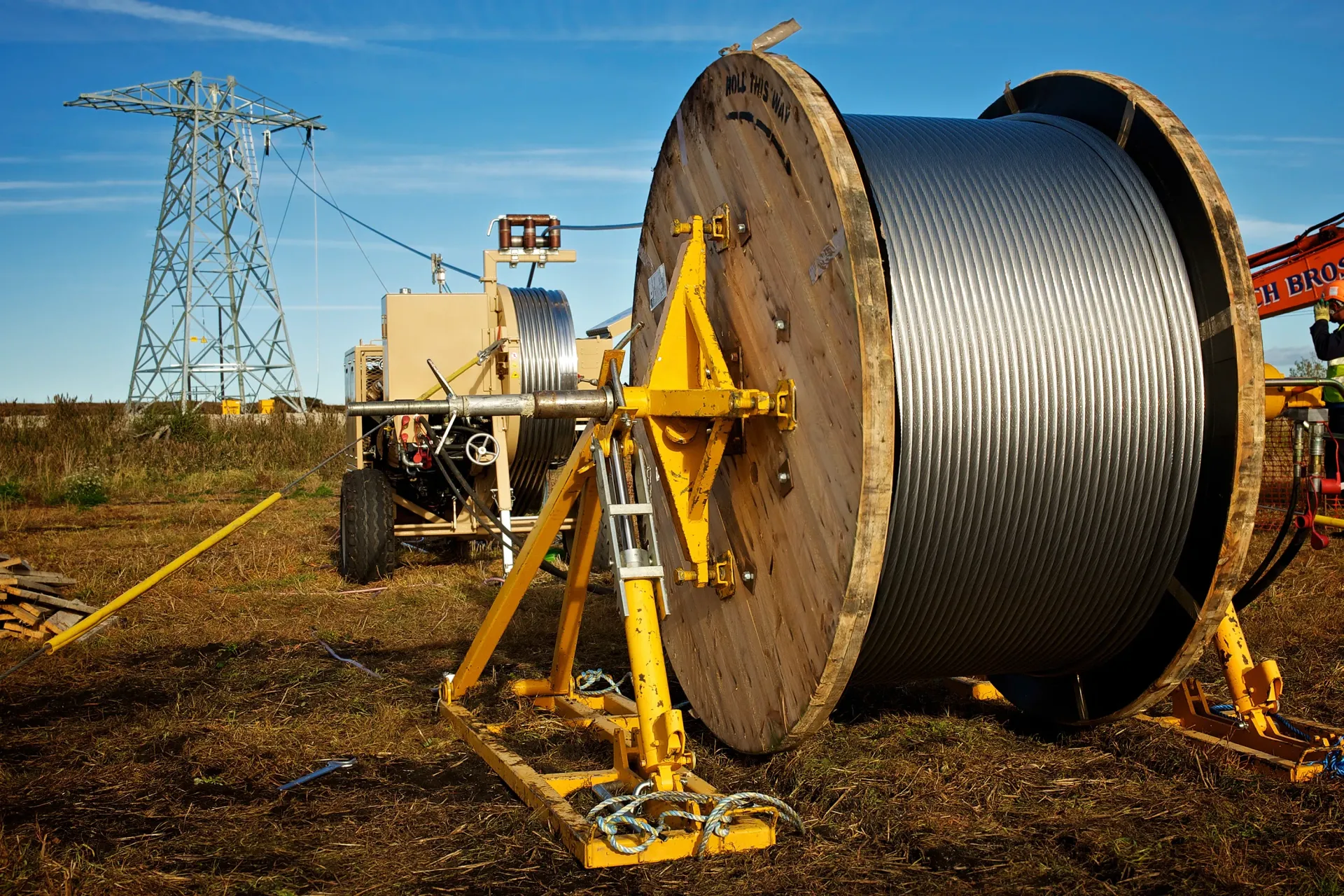 Large spool of black cable being unwound, with a power tower in the background on a sunny day.