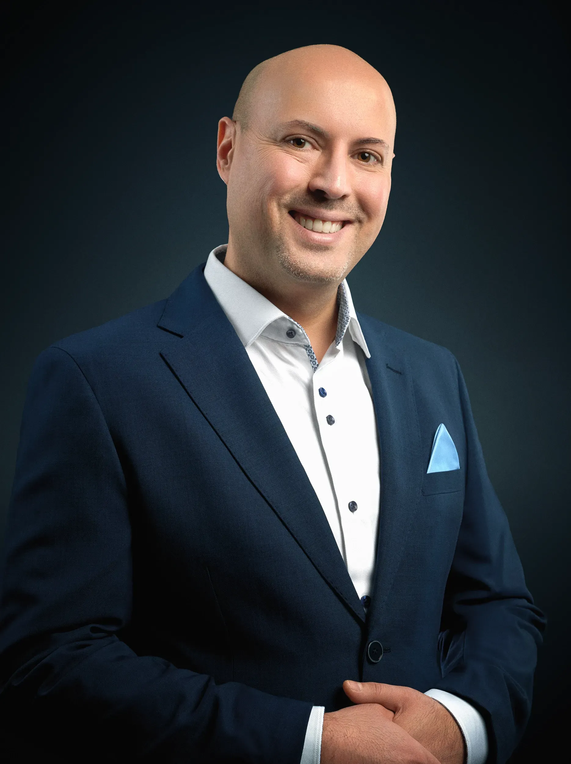 Smiling man in a blue suit with a light blue pocket square, against a dark blue background.
