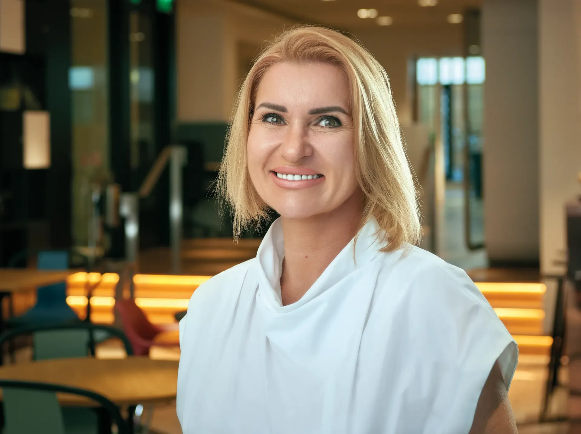 Woman with blonde hair smiles in a modern, brightly lit interior. She wears a white top.
