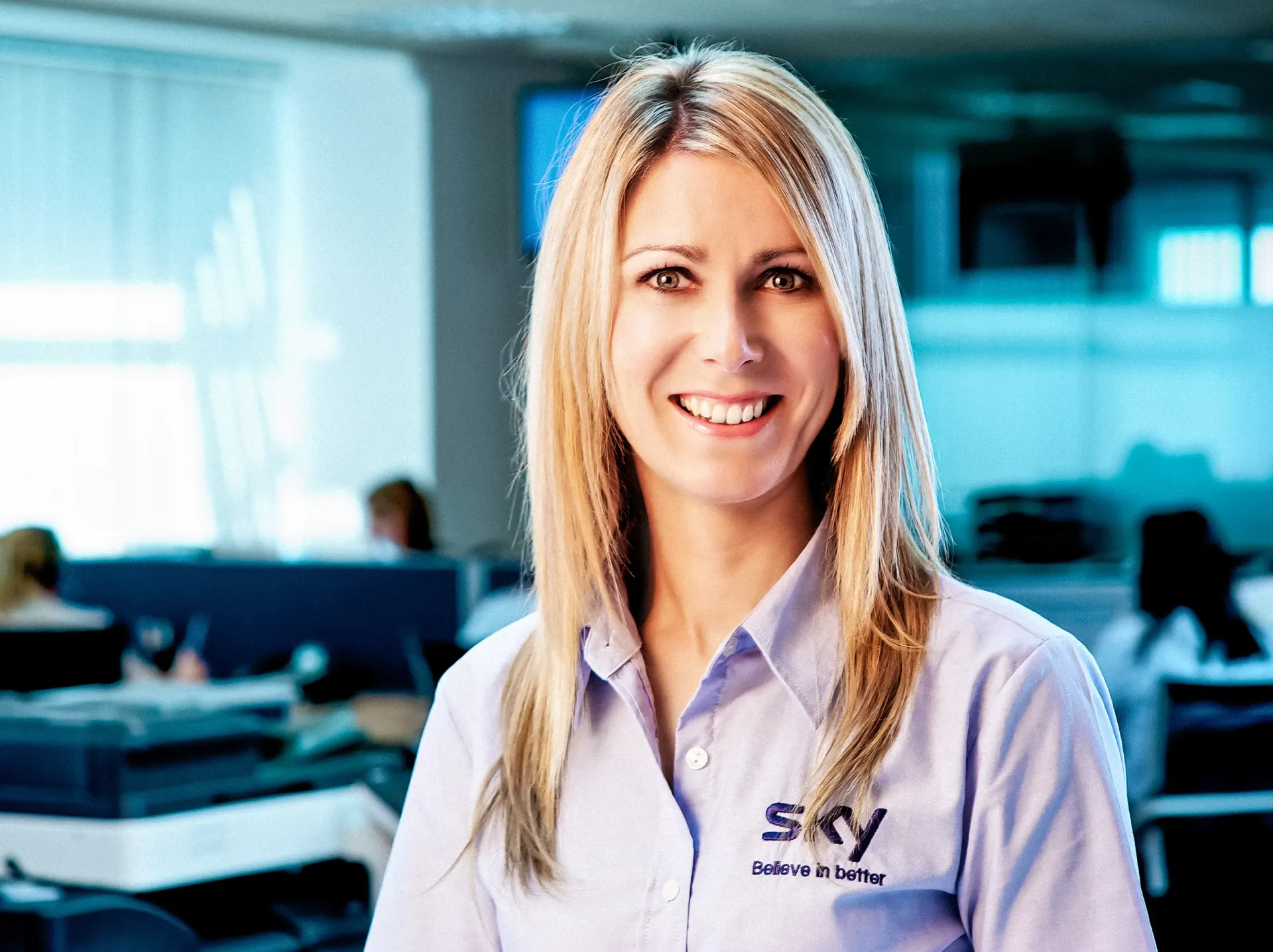 Woman with blonde hair wearing a Sky uniform shirt smiles in an office setting.