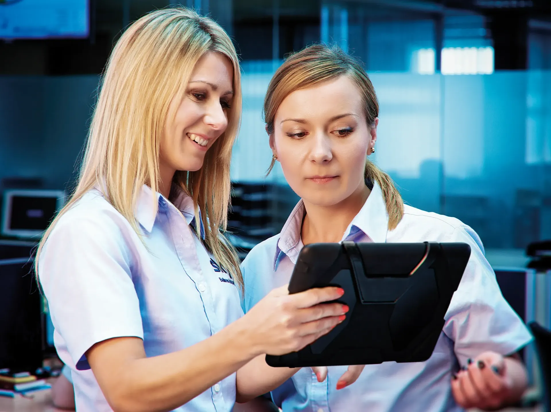 Two women in a modern office looking at a tablet, one smiling.