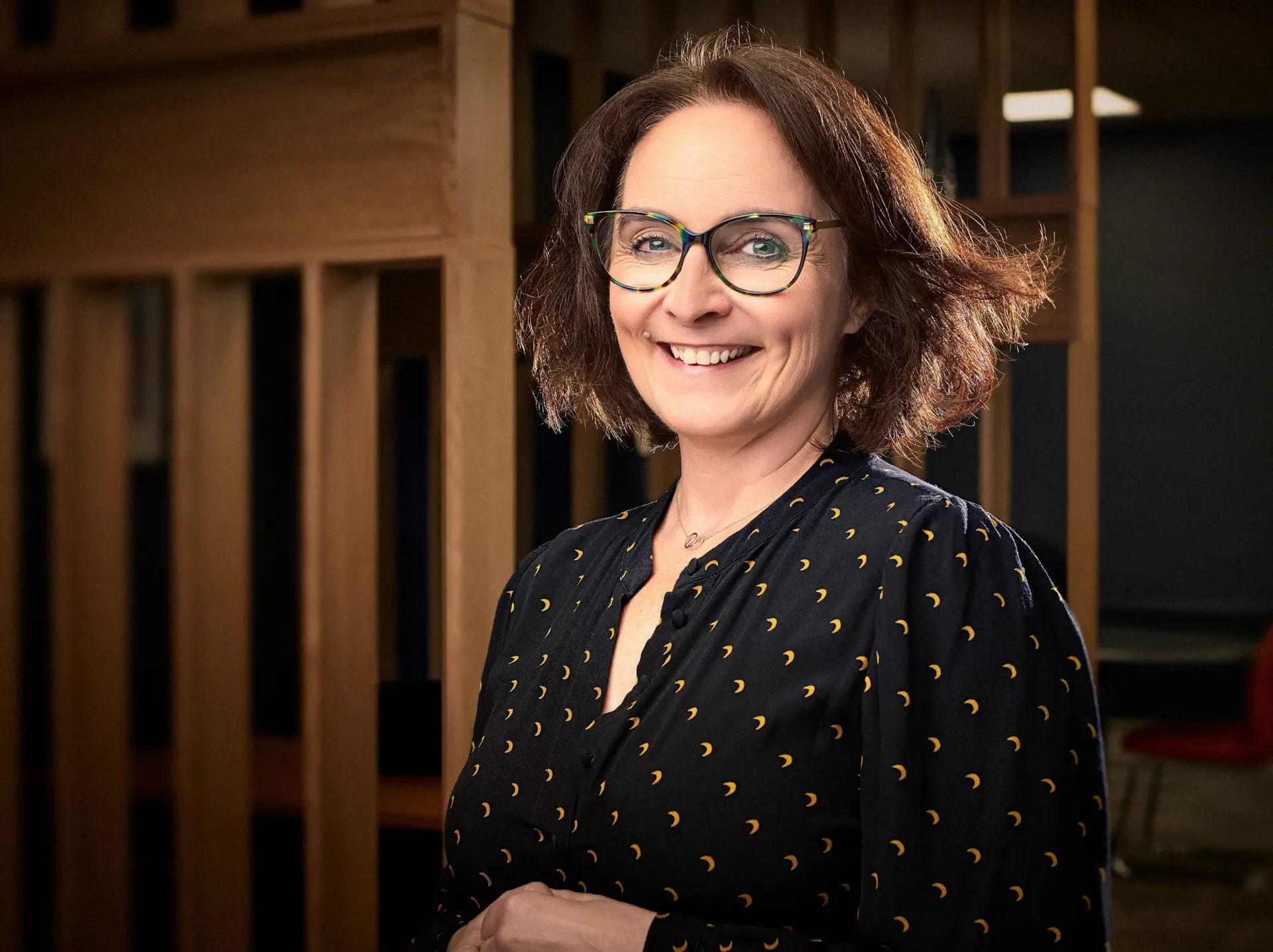 Woman with glasses smiles in a dark blue blouse, standing near a wooden structure.