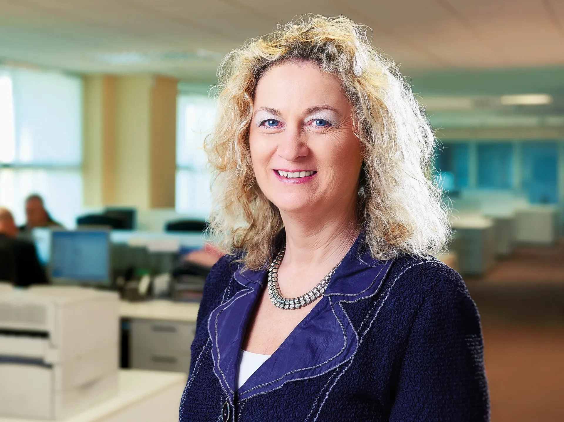 Woman in blazer smiles in an office, with open workspaces blurred in the background.