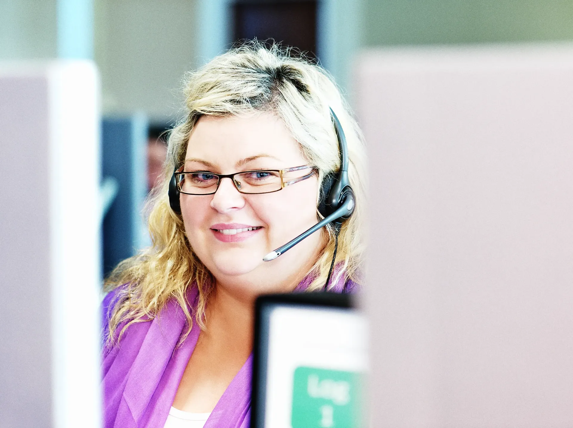 Woman with glasses and headset smiles while working at a computer in an office setting.