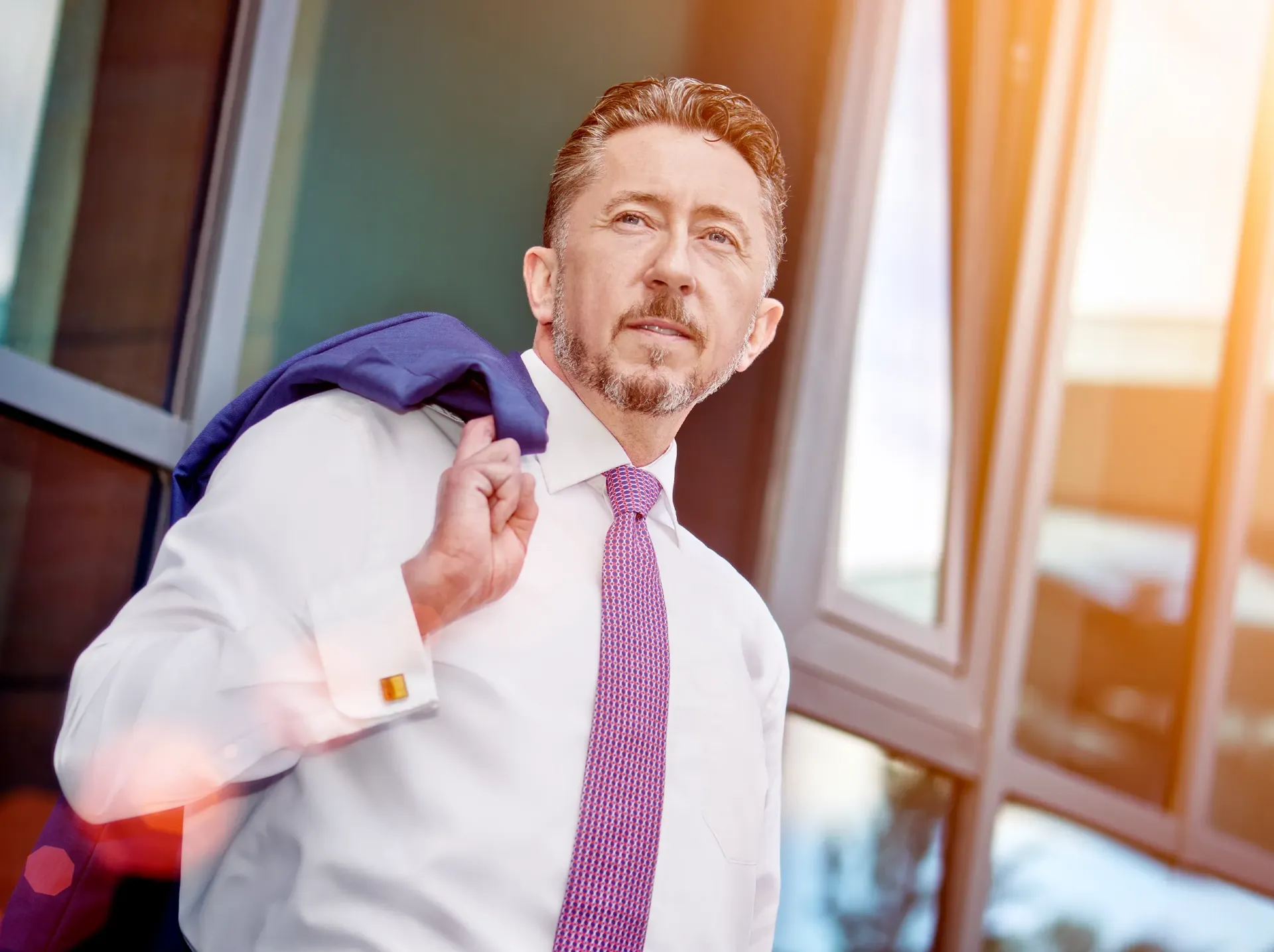 Man in white shirt and purple tie, jacket over his shoulder, near glass building in sunlight.