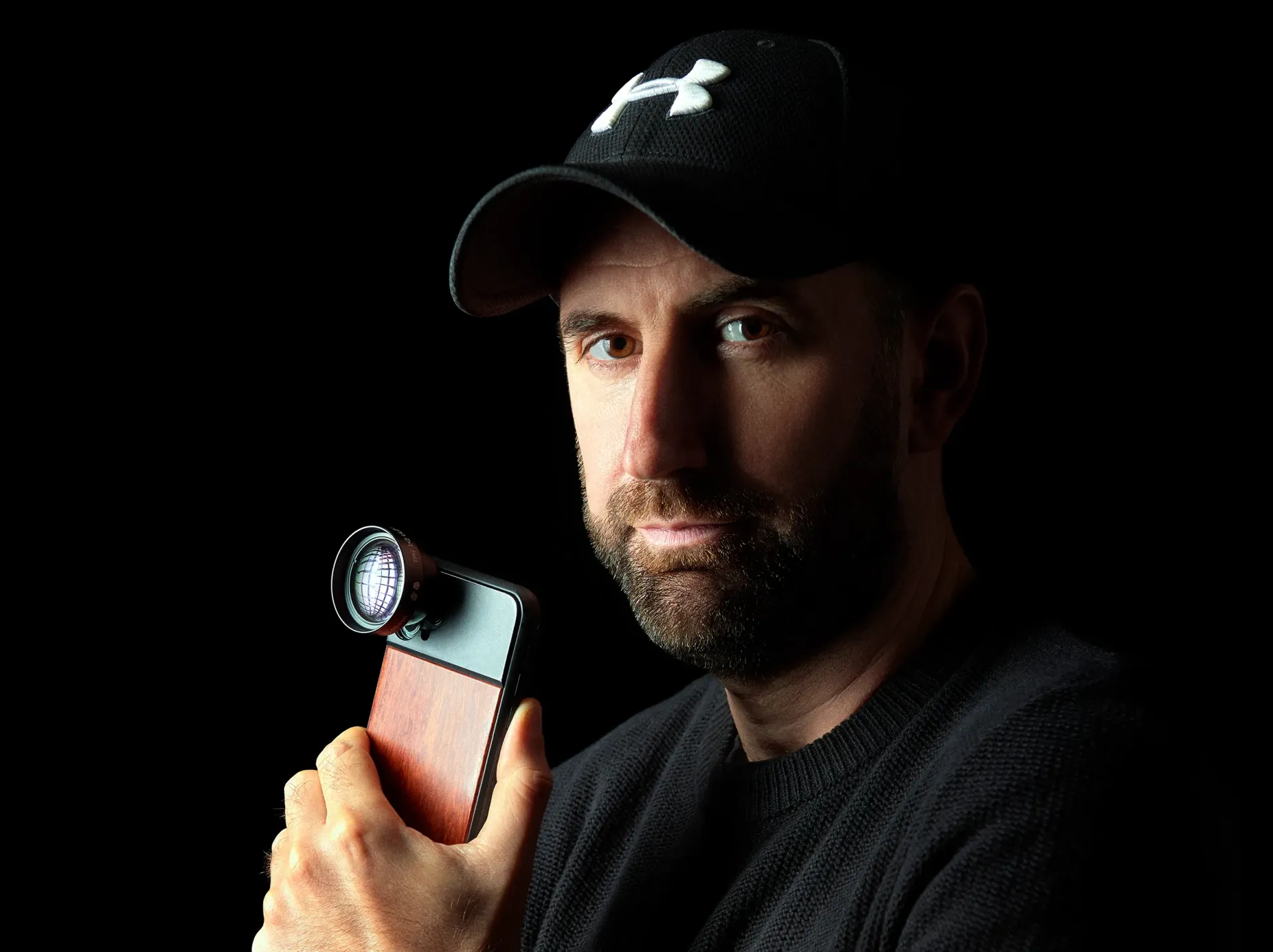 Man holding a smartphone with an attached lens, wearing a black cap and sweater, against a black backdrop.