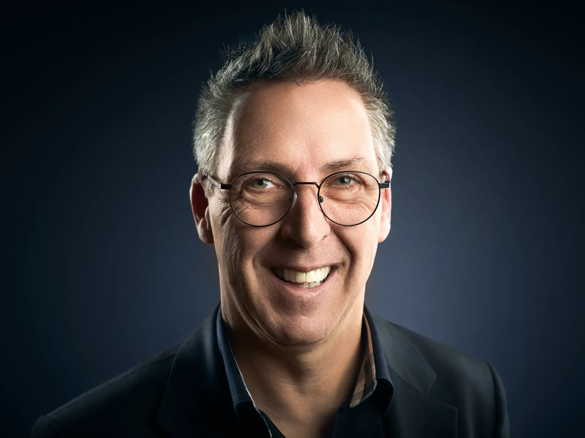 Smiling man wearing glasses and a dark blazer against a dark blue background.