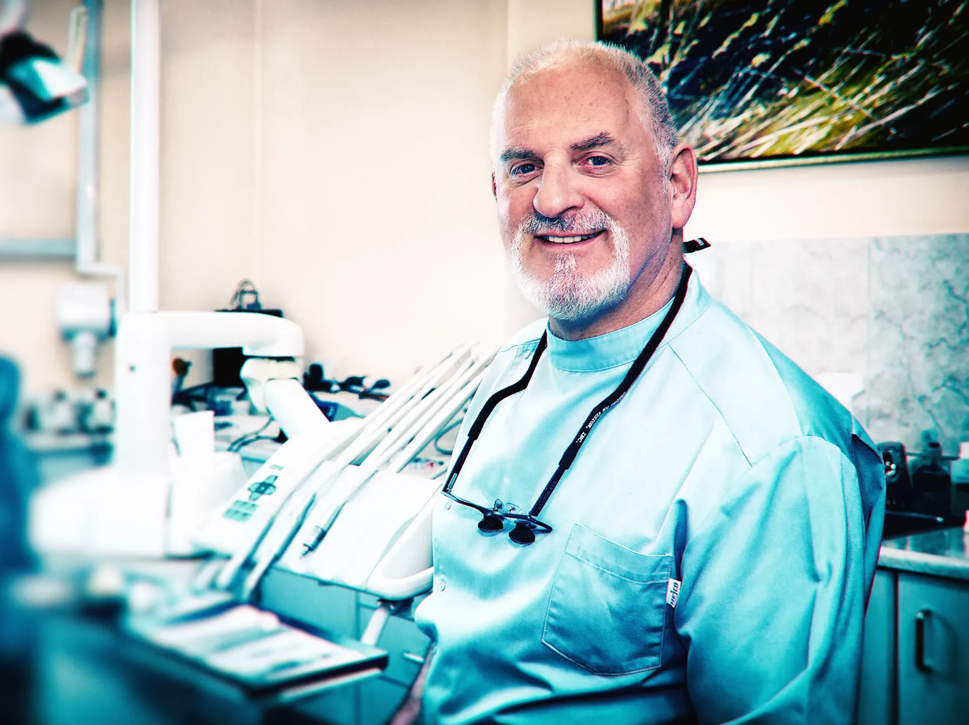Dentist smiling, wearing glasses and a light blue scrub, in a dental office.