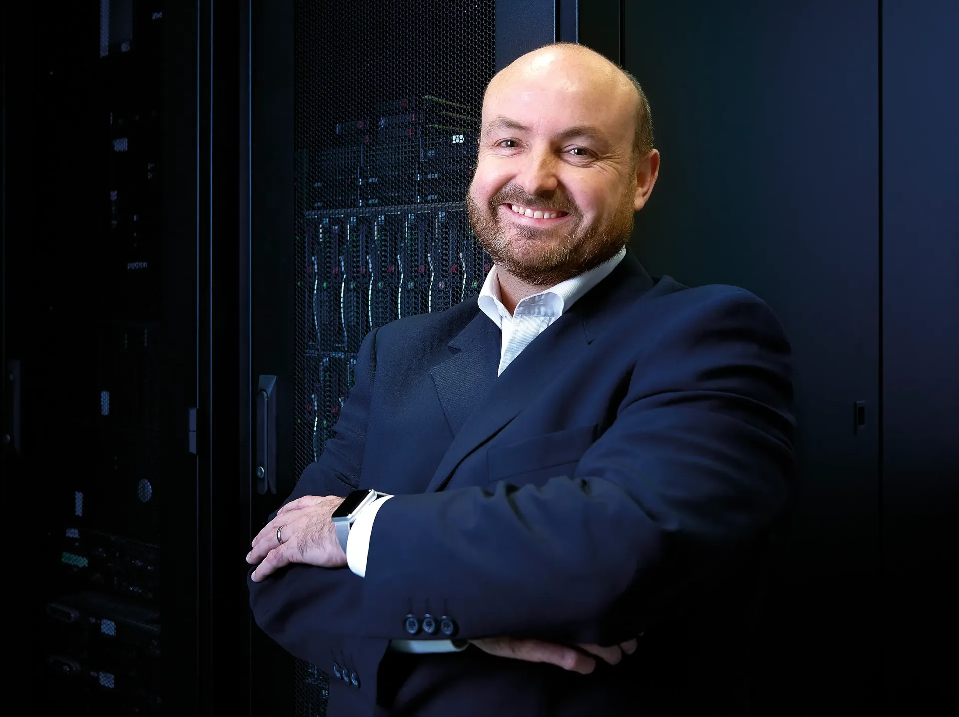 Man in a dark suit smiles with arms crossed, standing in a server room.