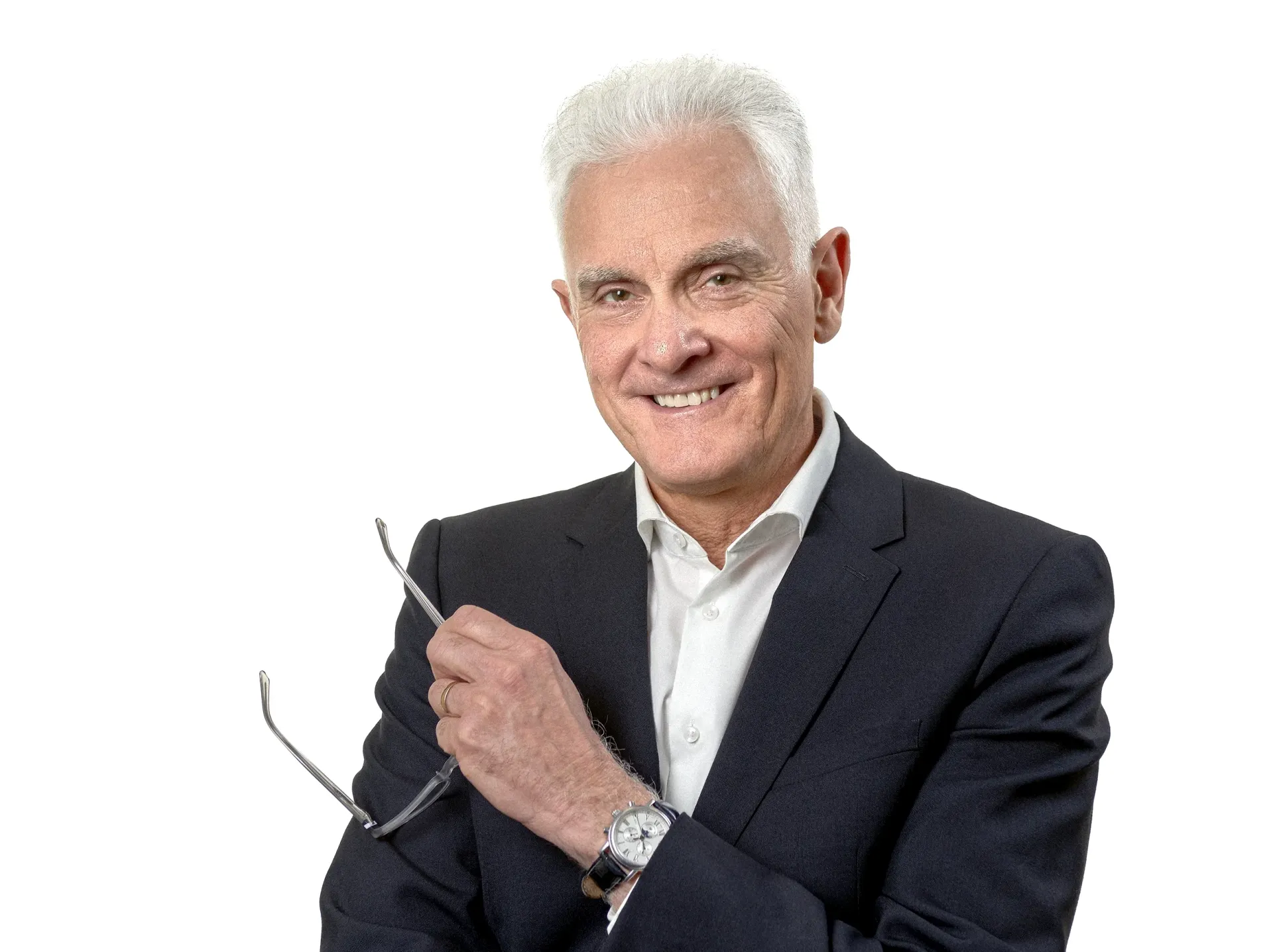 Man in a dark suit smiles, holding glasses, white hair, against a white background.