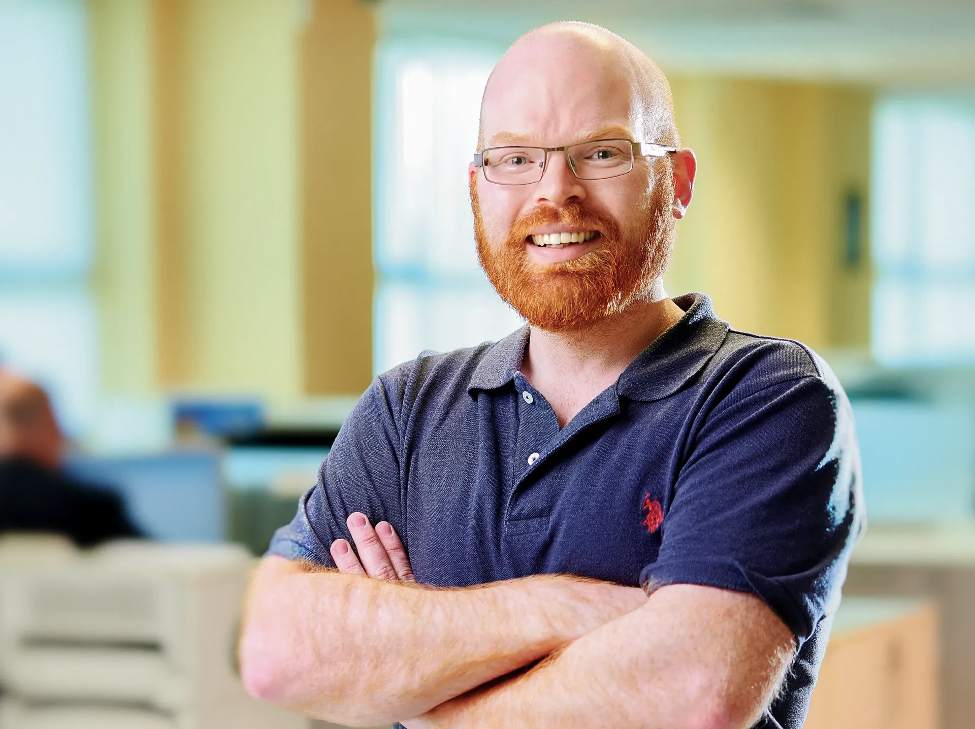 Bald man with red beard and glasses smiles, arms crossed, in a blurred office setting.