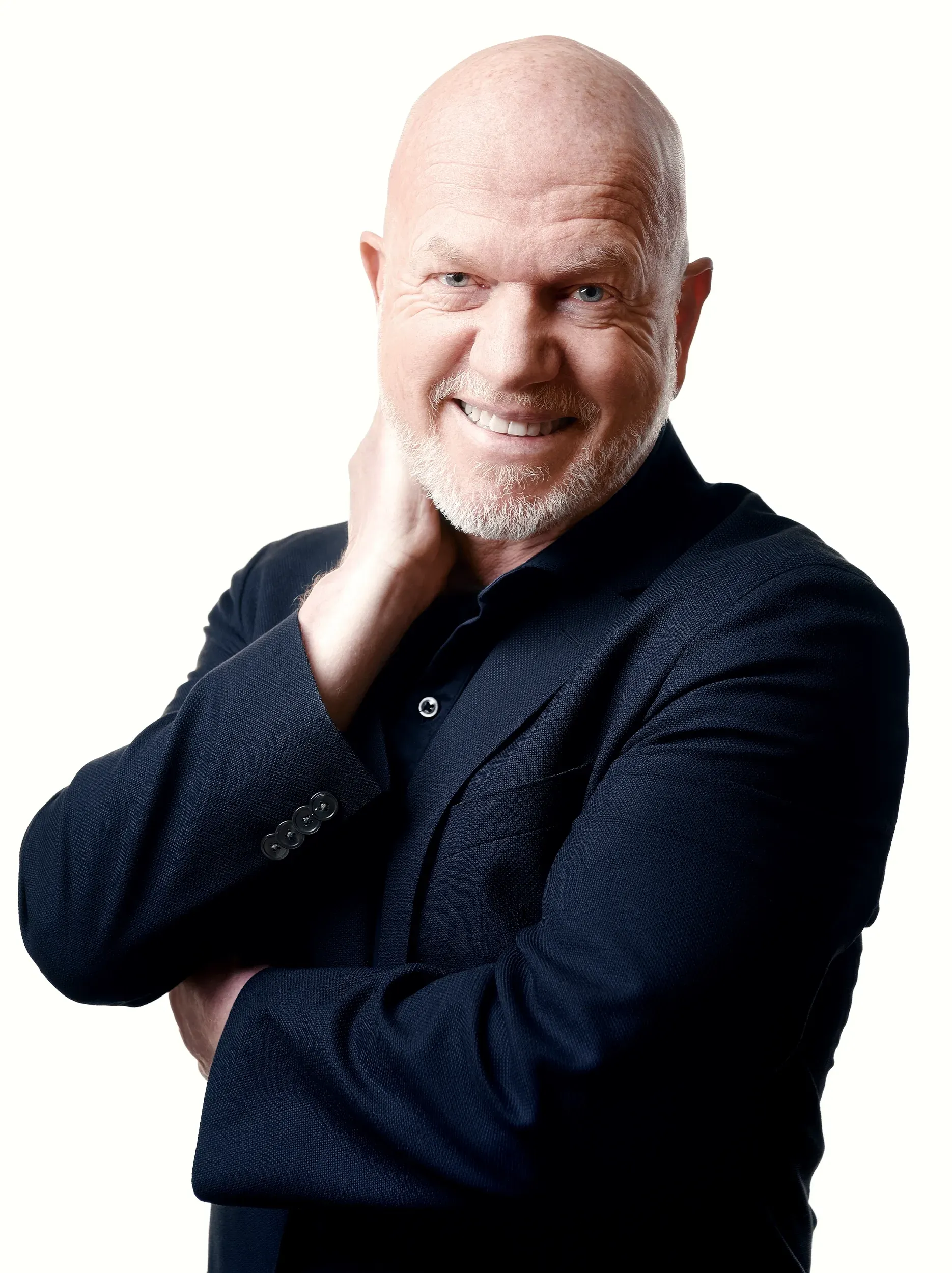 Bald man in black blazer smiles, hand at neck, leaning.