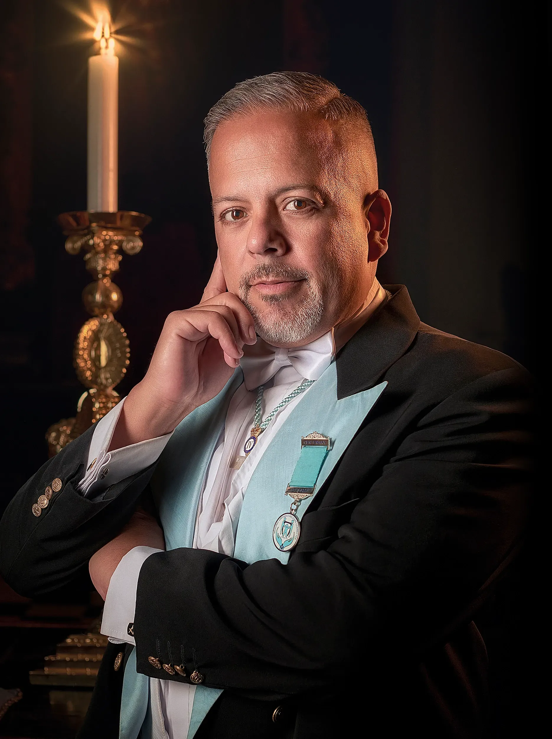 A man in a formal black suit with a light blue sash and an award pin poses beside a lit candle in a dim, elegant setting.