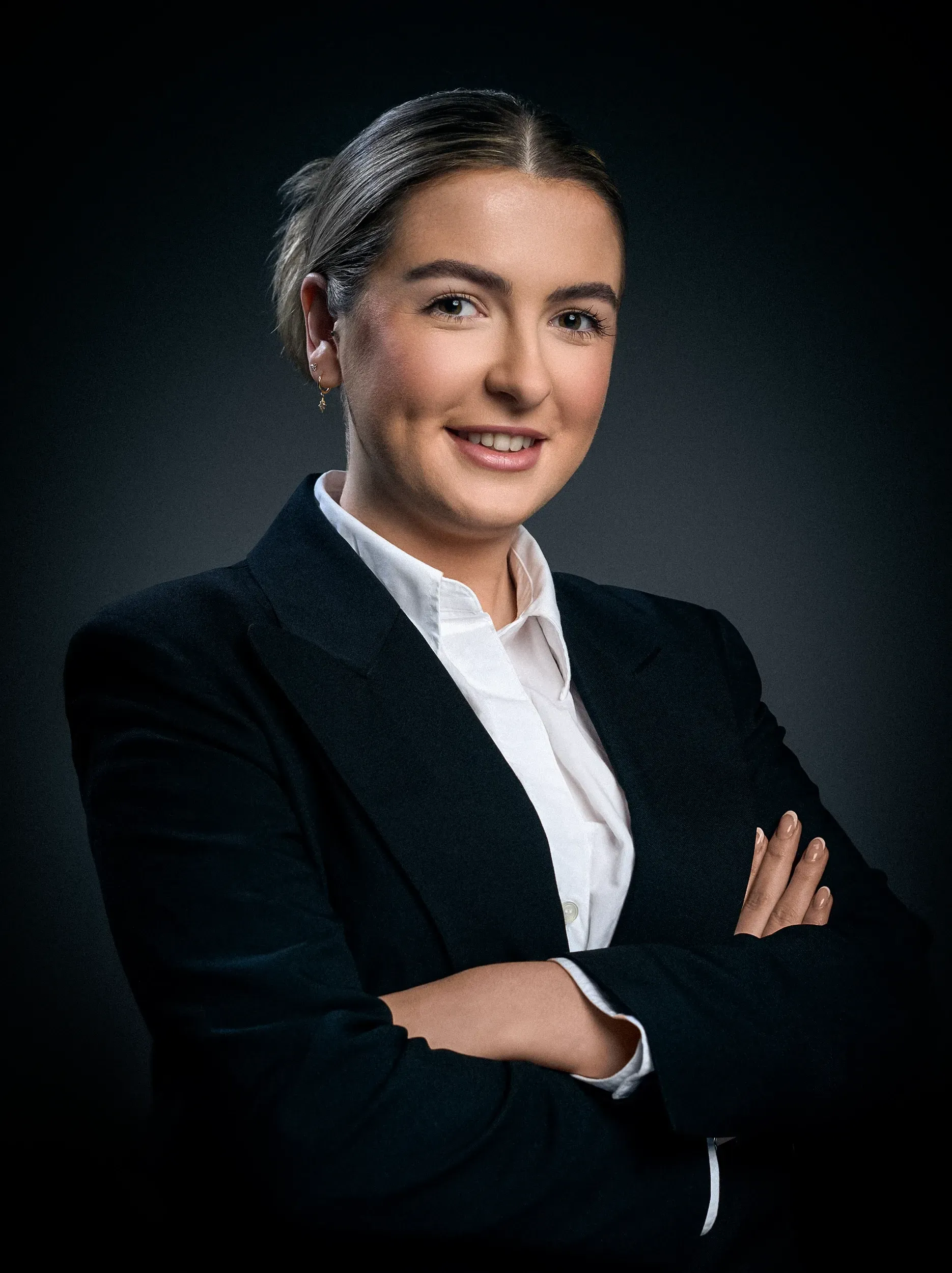 Woman in suit with arms crossed, smiling, against a dark gray background.