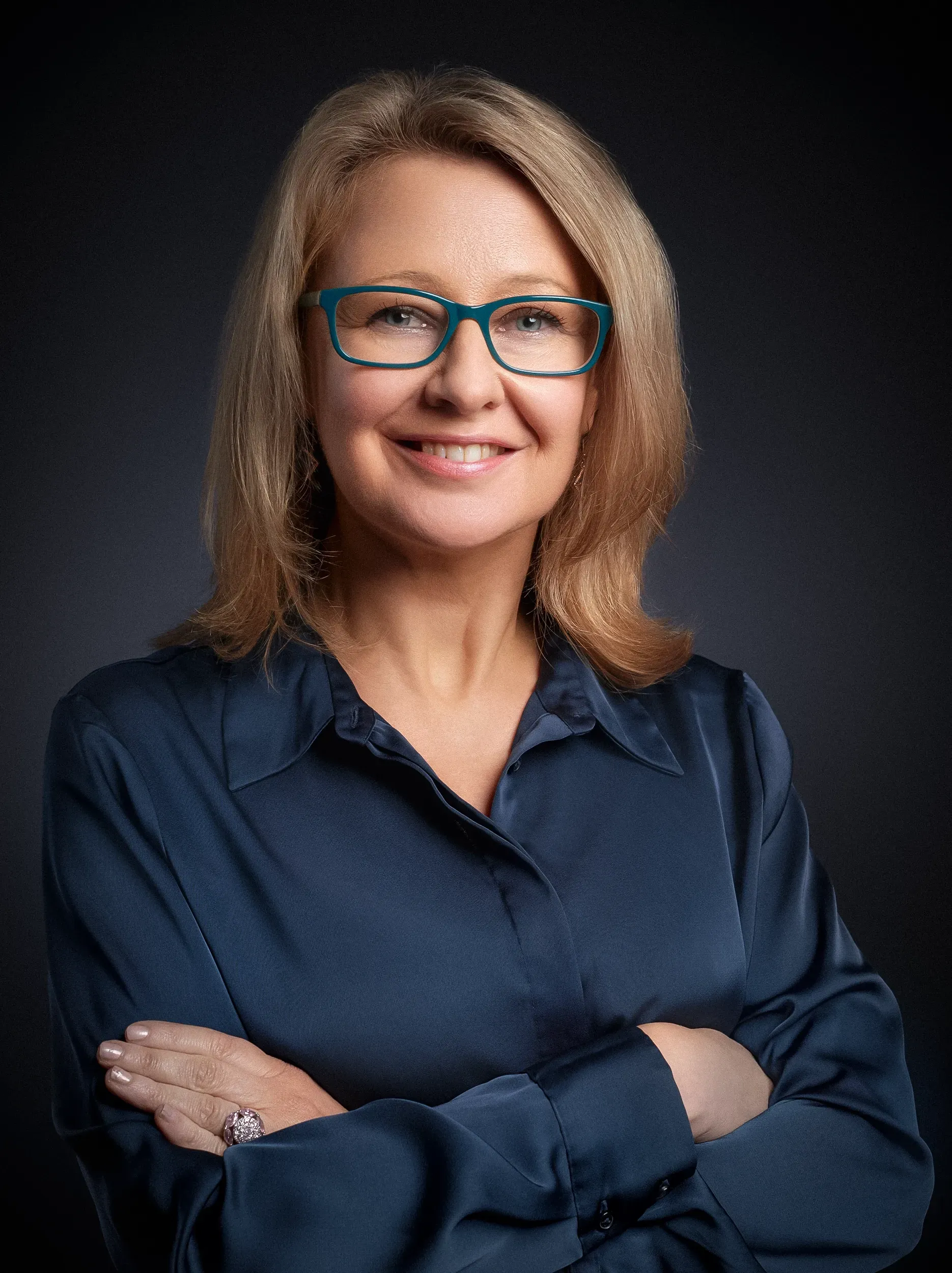 Woman in teal glasses and blue blouse smiles, arms crossed against a dark gray background.