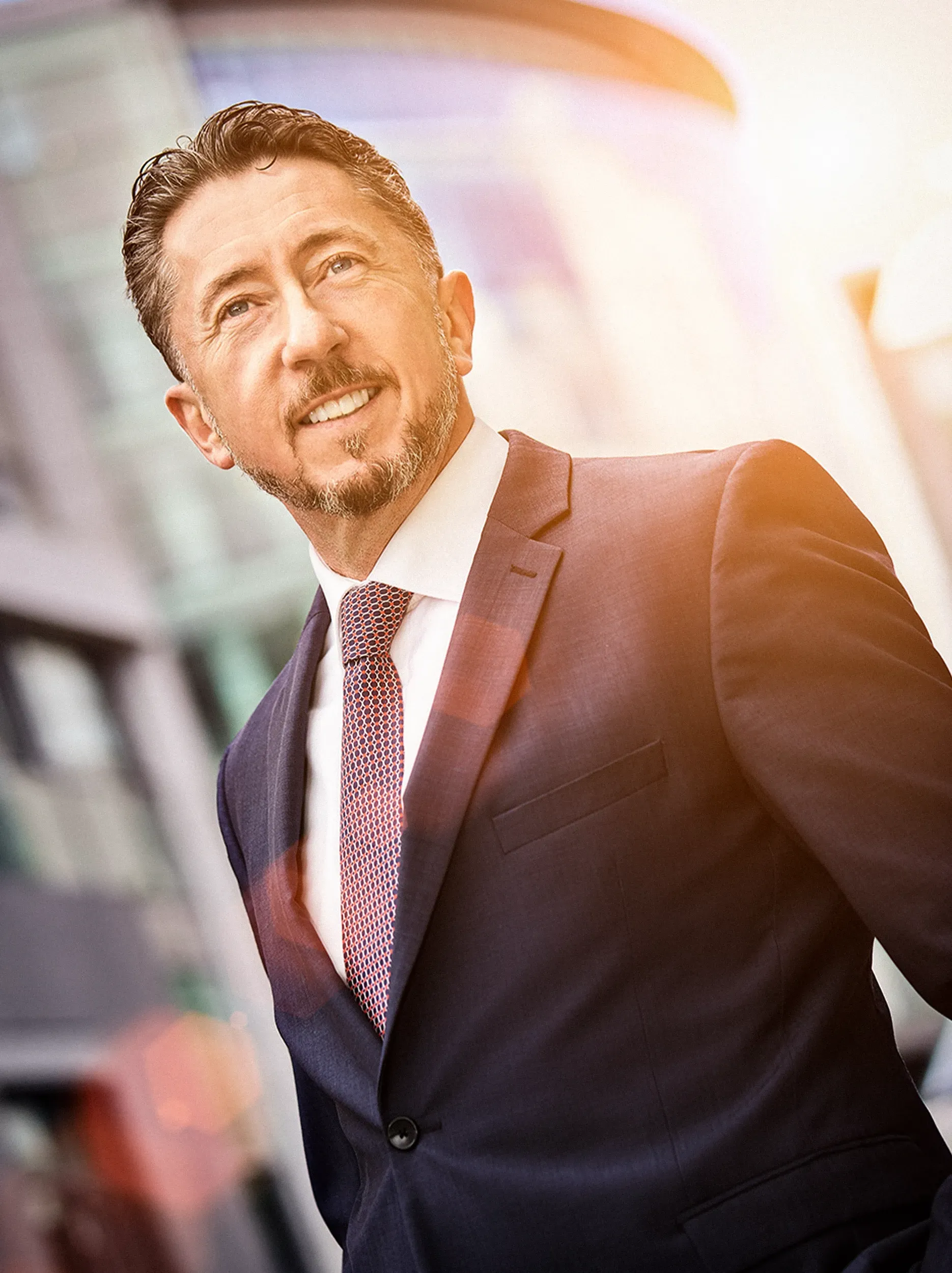 Man in a suit smiles, outdoors in front of a modern building.