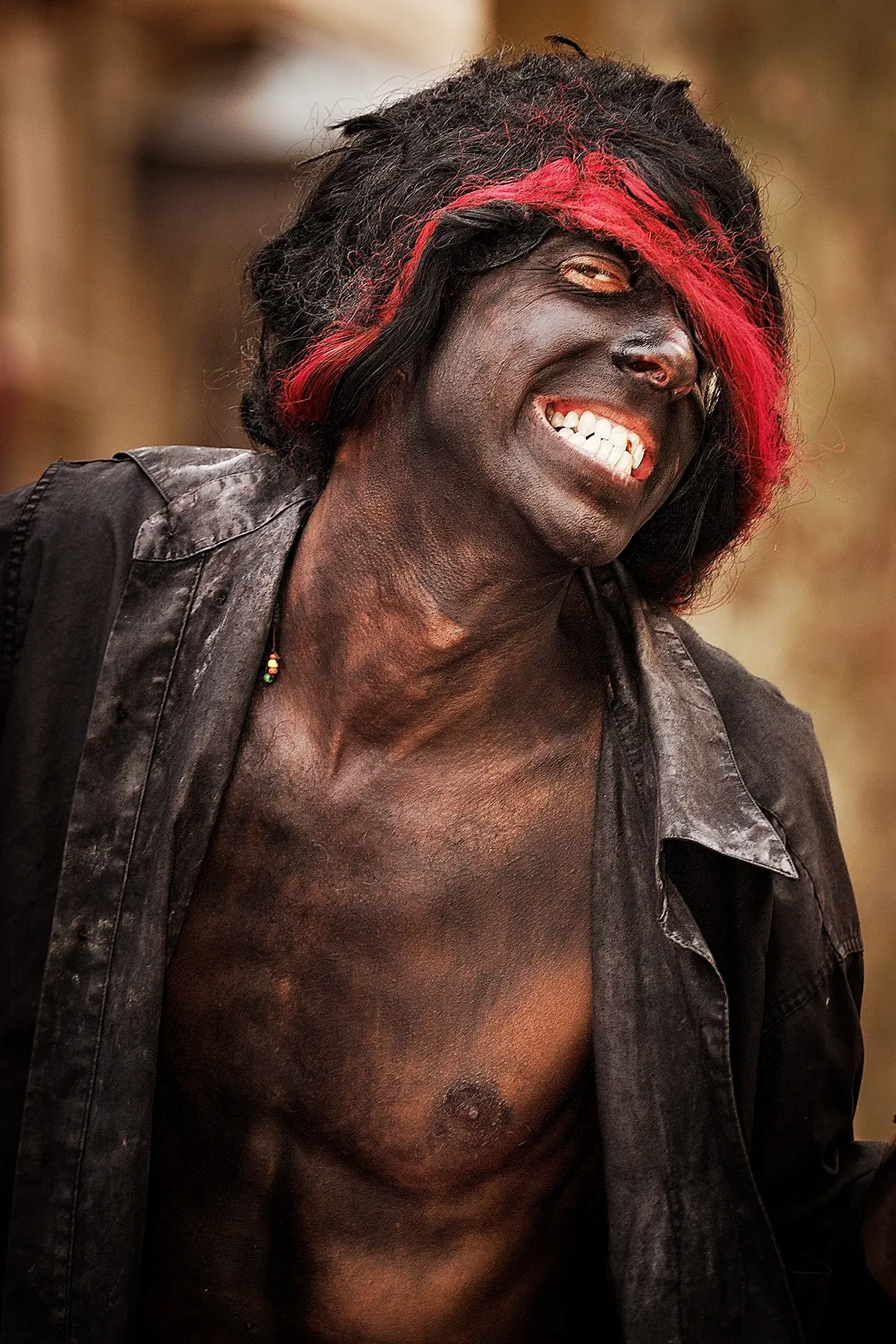 Person with dark face and chest, black hair with red streak, smiling, wearing open black shirt.