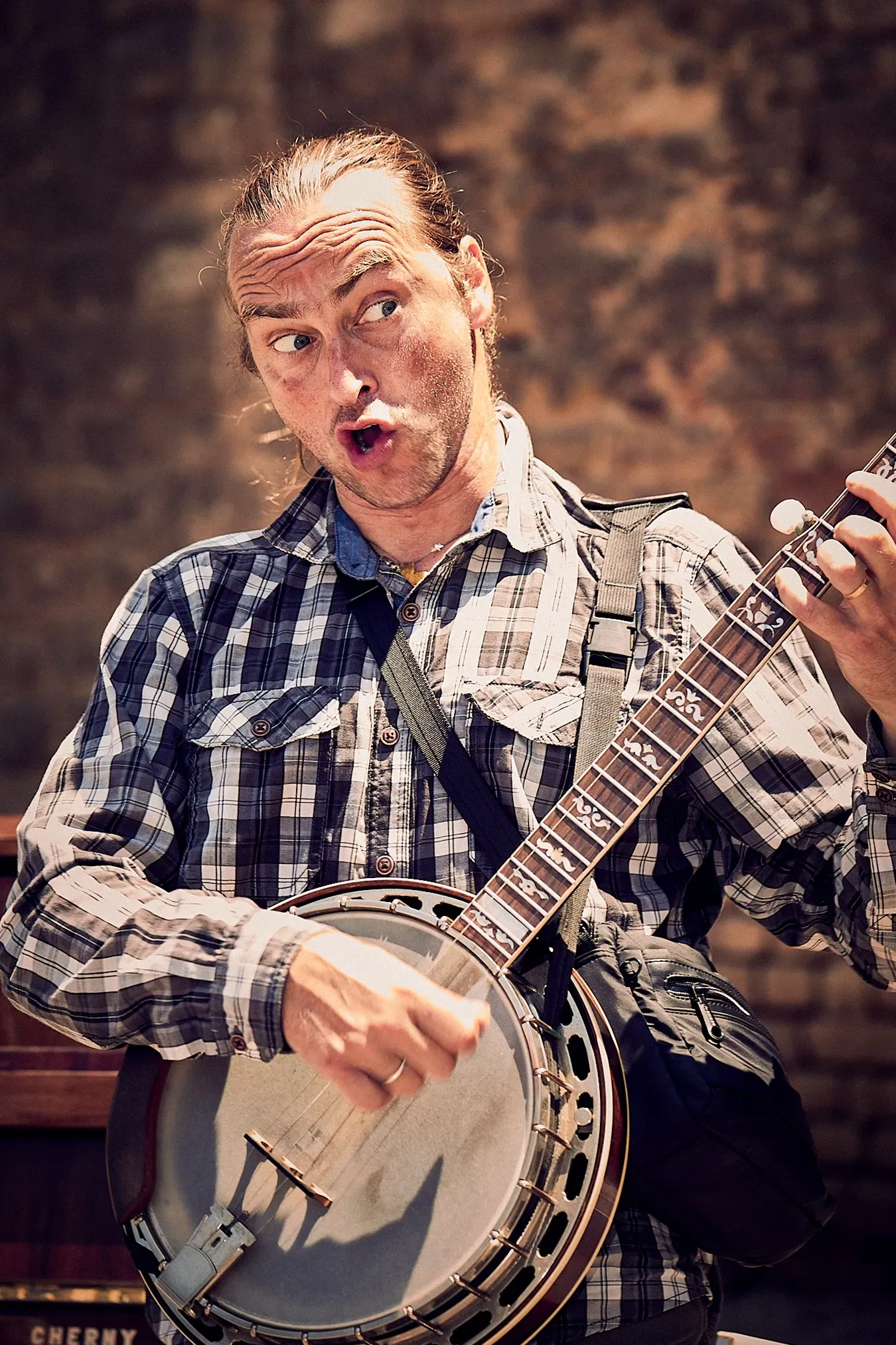 Man playing a banjo, surprised expression, outdoors with brick wall background.