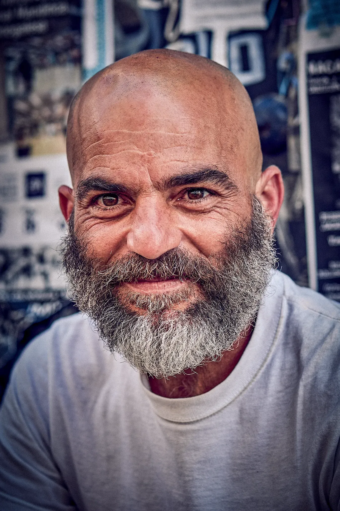 Bald man with a salt-and-pepper beard, smiling slightly. Wearing a white t-shirt, background has posters.