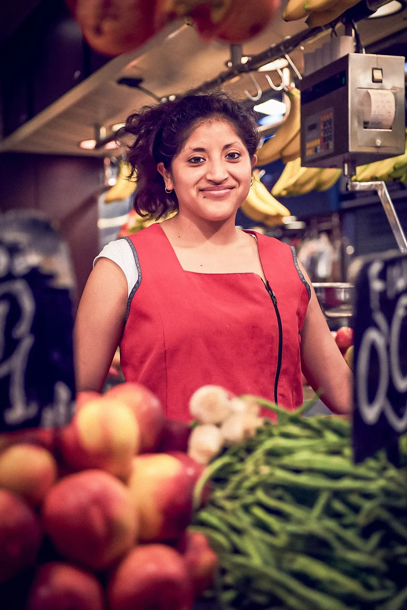 Woman in red apron smiles behind a produce stand. Fruits and vegetables are in front and a scale is behind her.