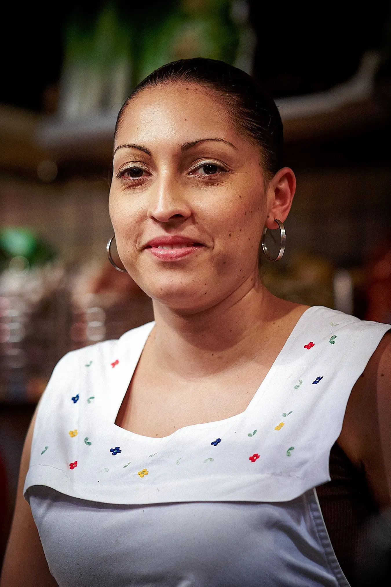 Woman wearing white apron, smiling, with hoop earrings, in a restaurant setting.