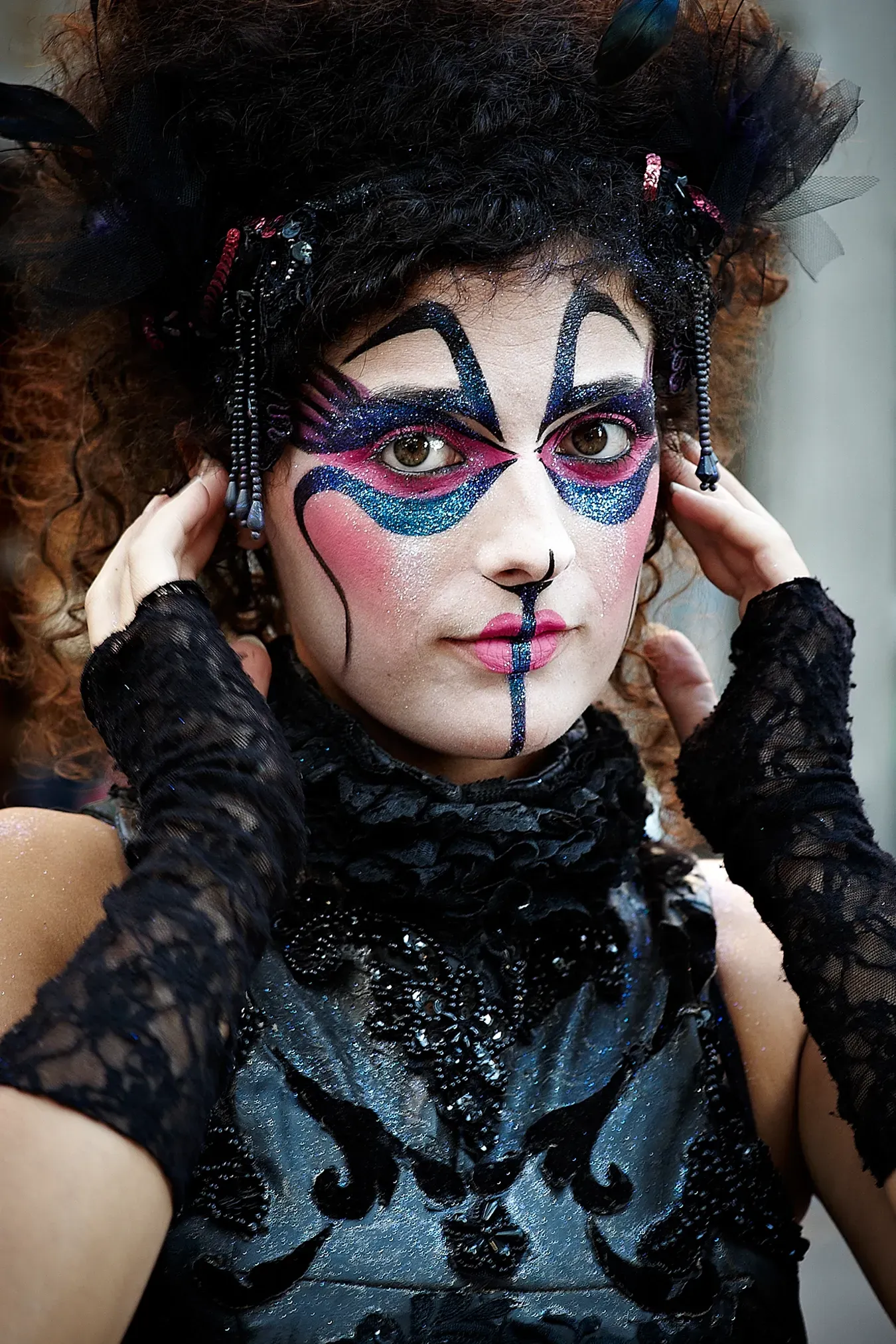 Woman in ornate black costume with dramatic face paint, touching her temples.