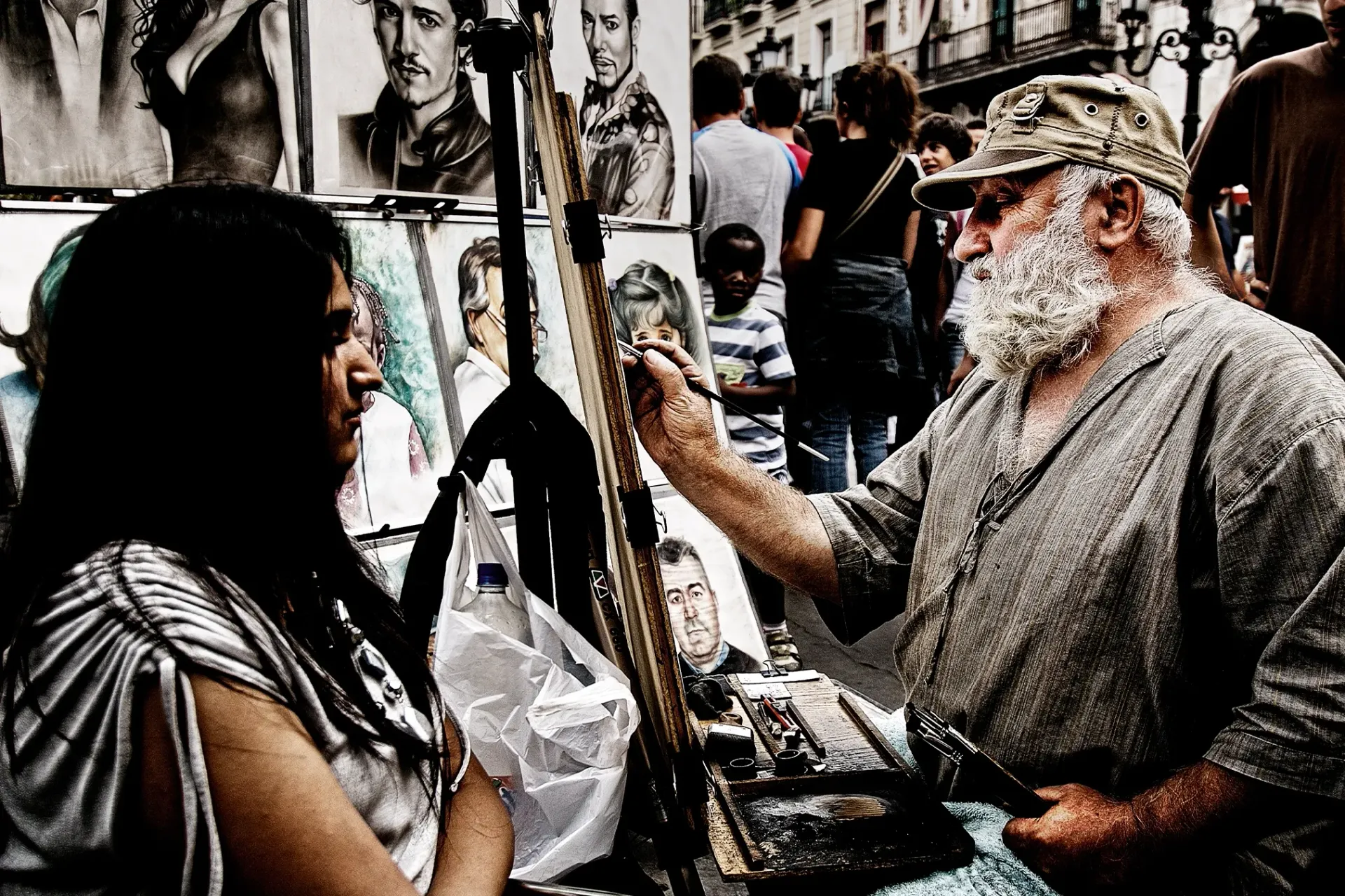 Street artist drawing a portrait of a person. Artist with white beard, drawing on easel. Bystanders in background.