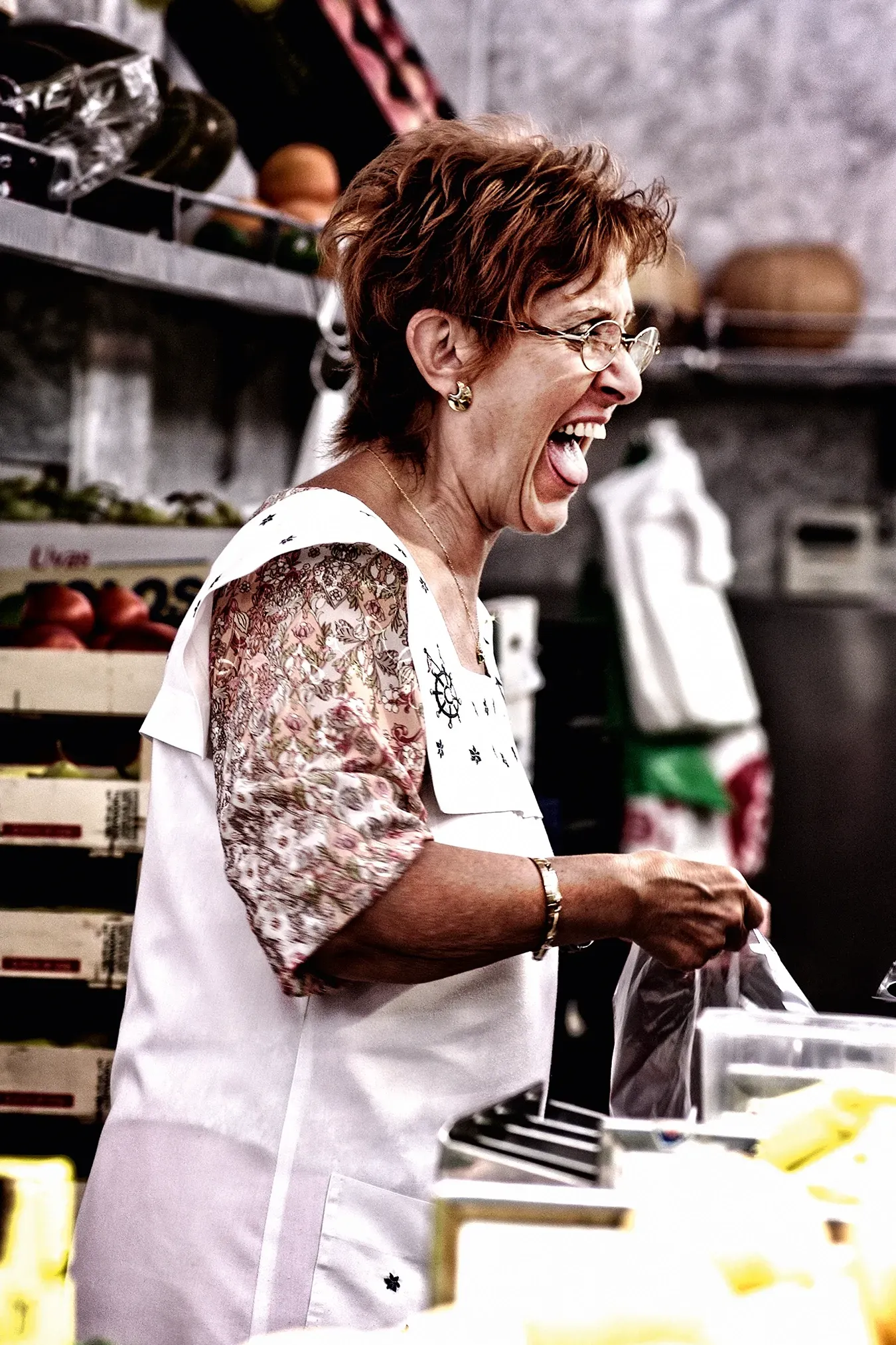 Woman laughing while working at a market counter, wearing glasses and a floral-sleeved top.