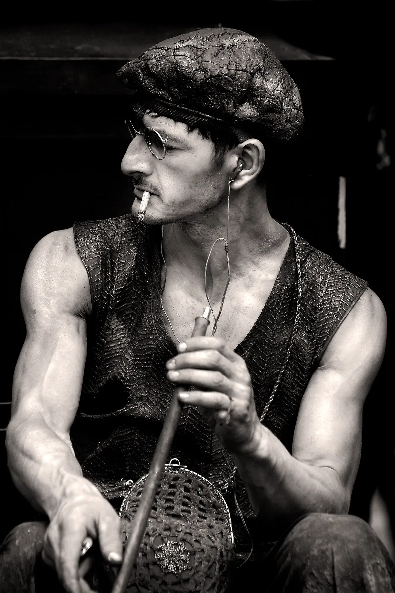 Man with beret, vest, cigarette, holding glassblowing tool, listening to music through earphones.