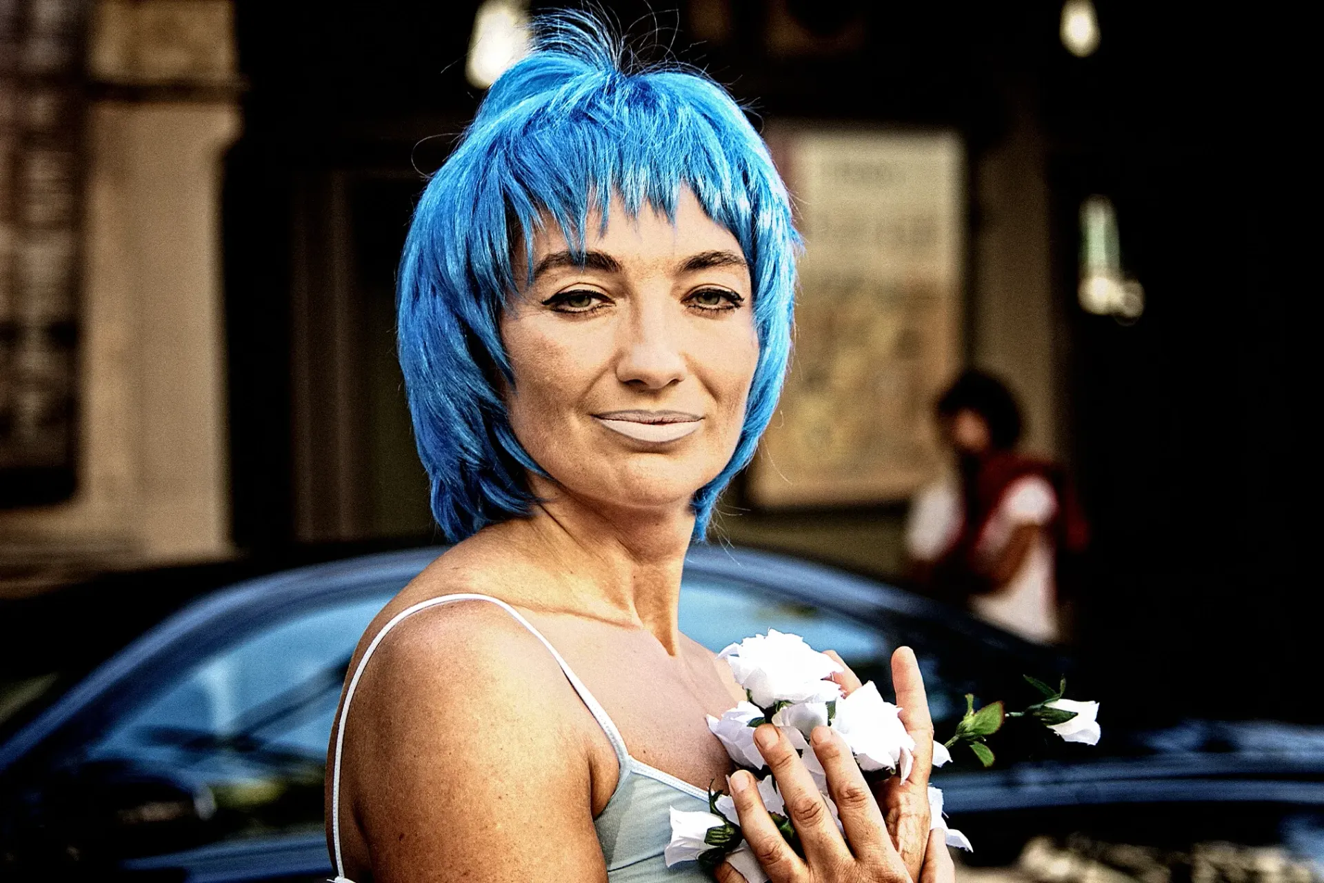 Woman with blue hair holds white flowers; wears a light blue dress. Building and car in background.