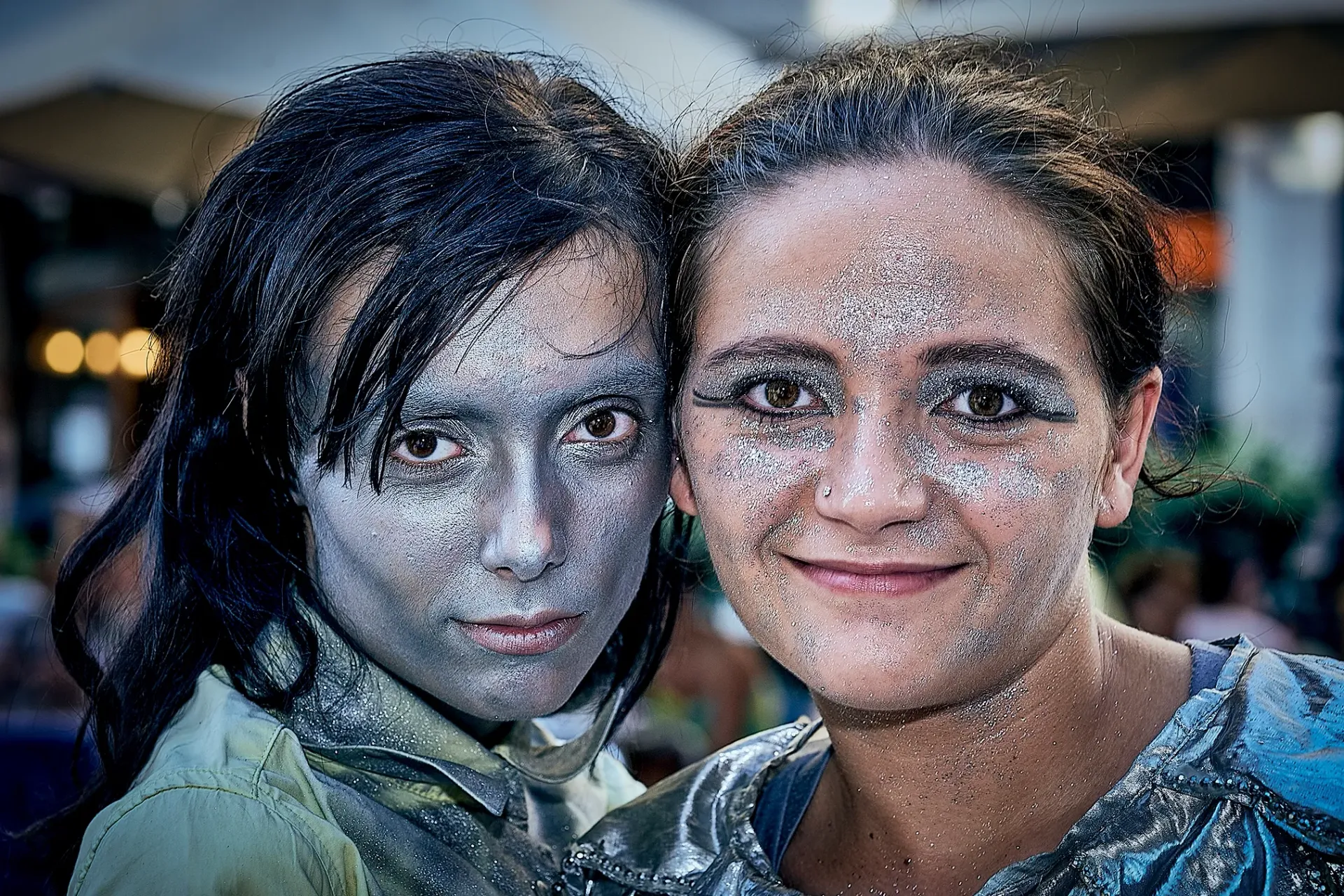 Two people with silver face paint, smiling, outside.