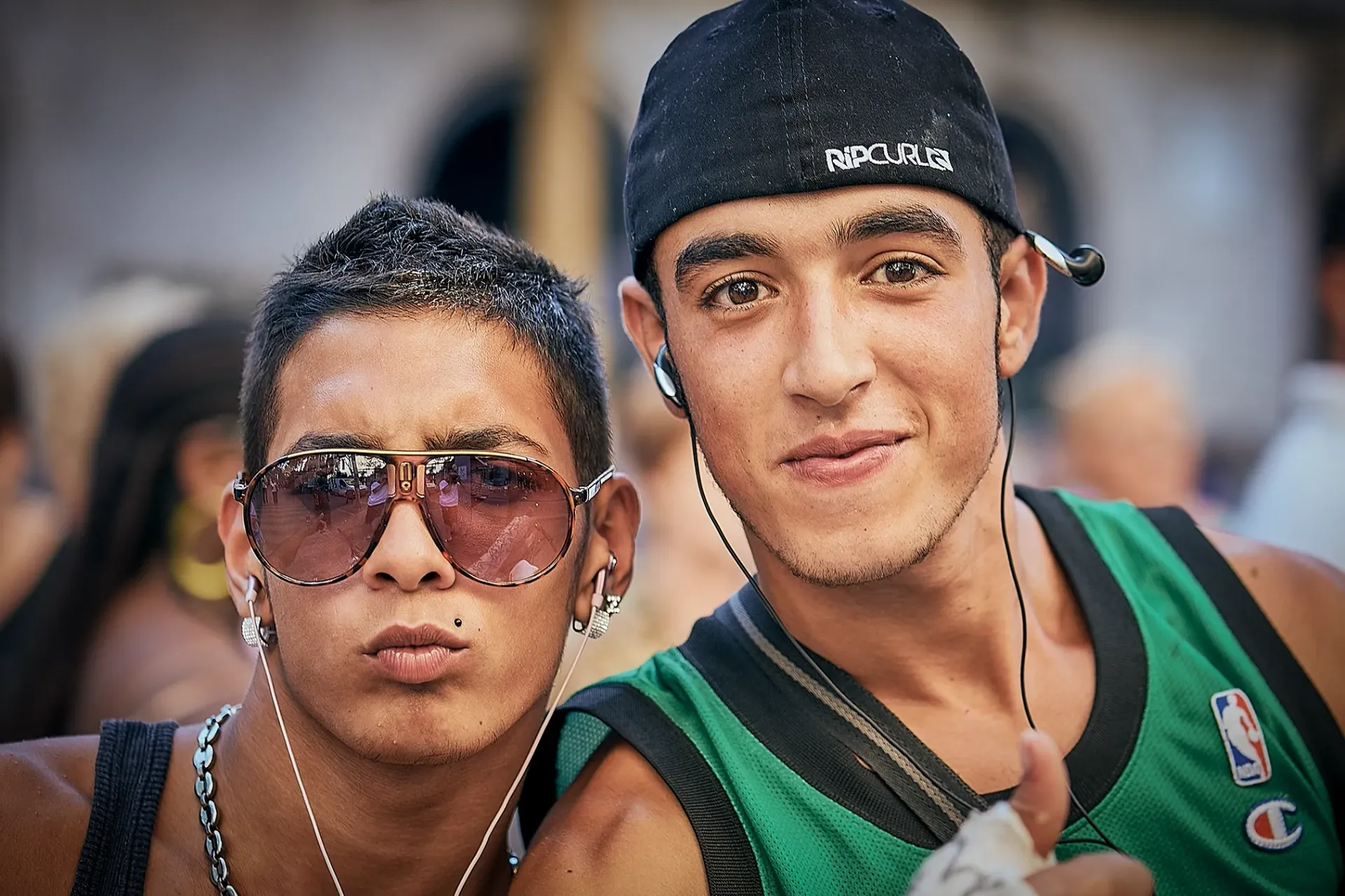 Two young men posing, one with sunglasses, the other with a hat and earbuds. Outdoors, sunny.