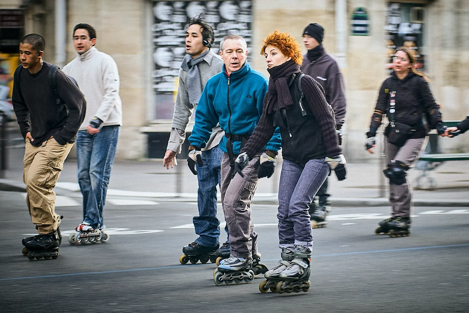 People inline skating on a street. Some are wearing protective gear. Buildings in the background.
