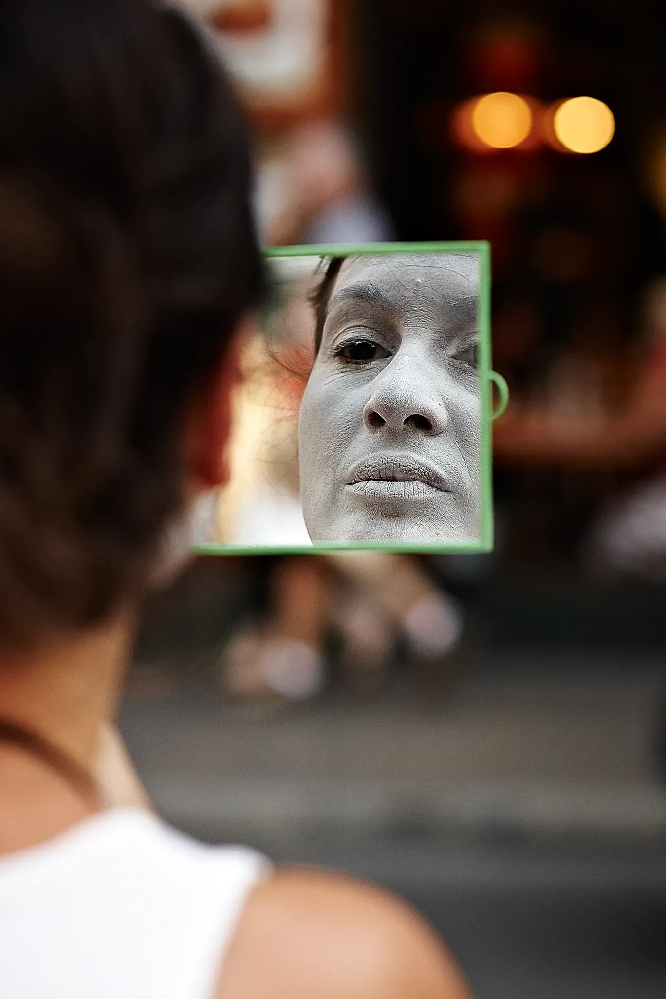 Woman holding a small mirror, face covered in white. Focus on the reflection, outdoors.