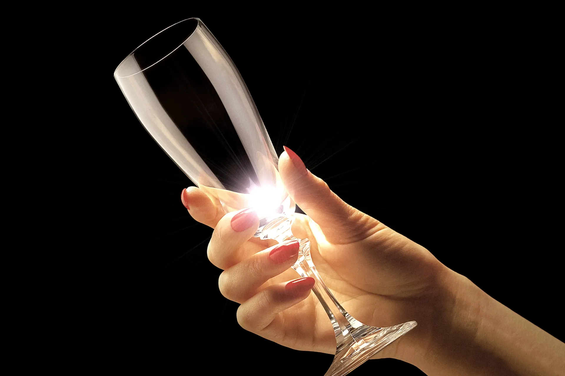 Hand holding a champagne flute against a black background, light illuminating the glass.