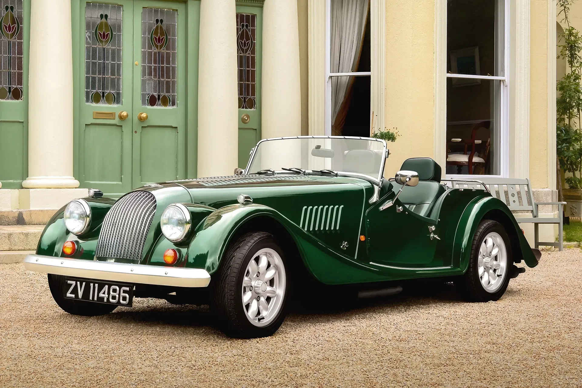 Green Morgan sports car parked in front of a light-yellow building with green doors.