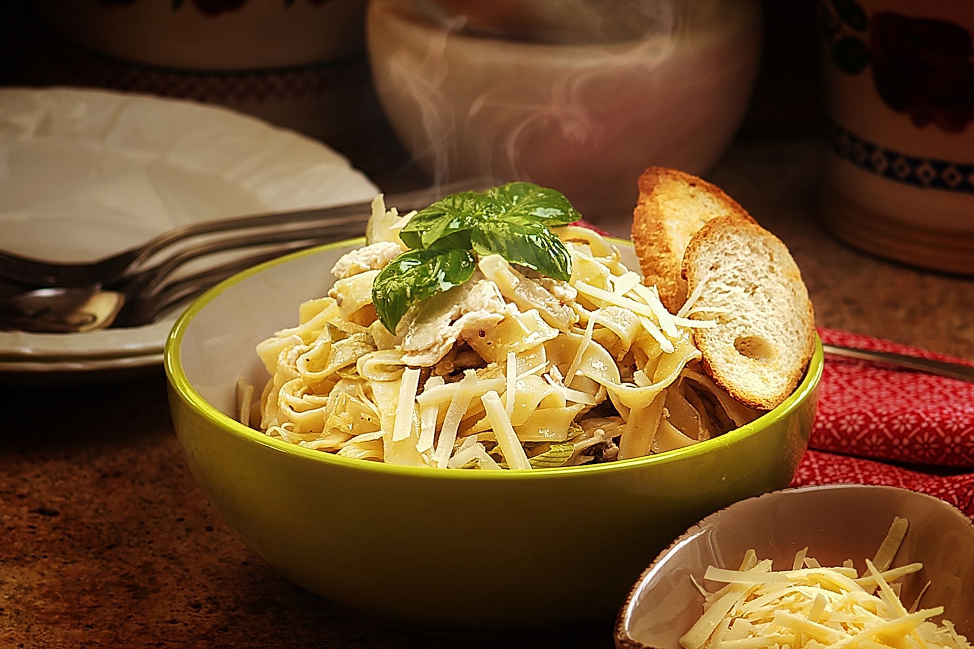Pasta dish in a green bowl with bread, cheese, and basil, steam rising.