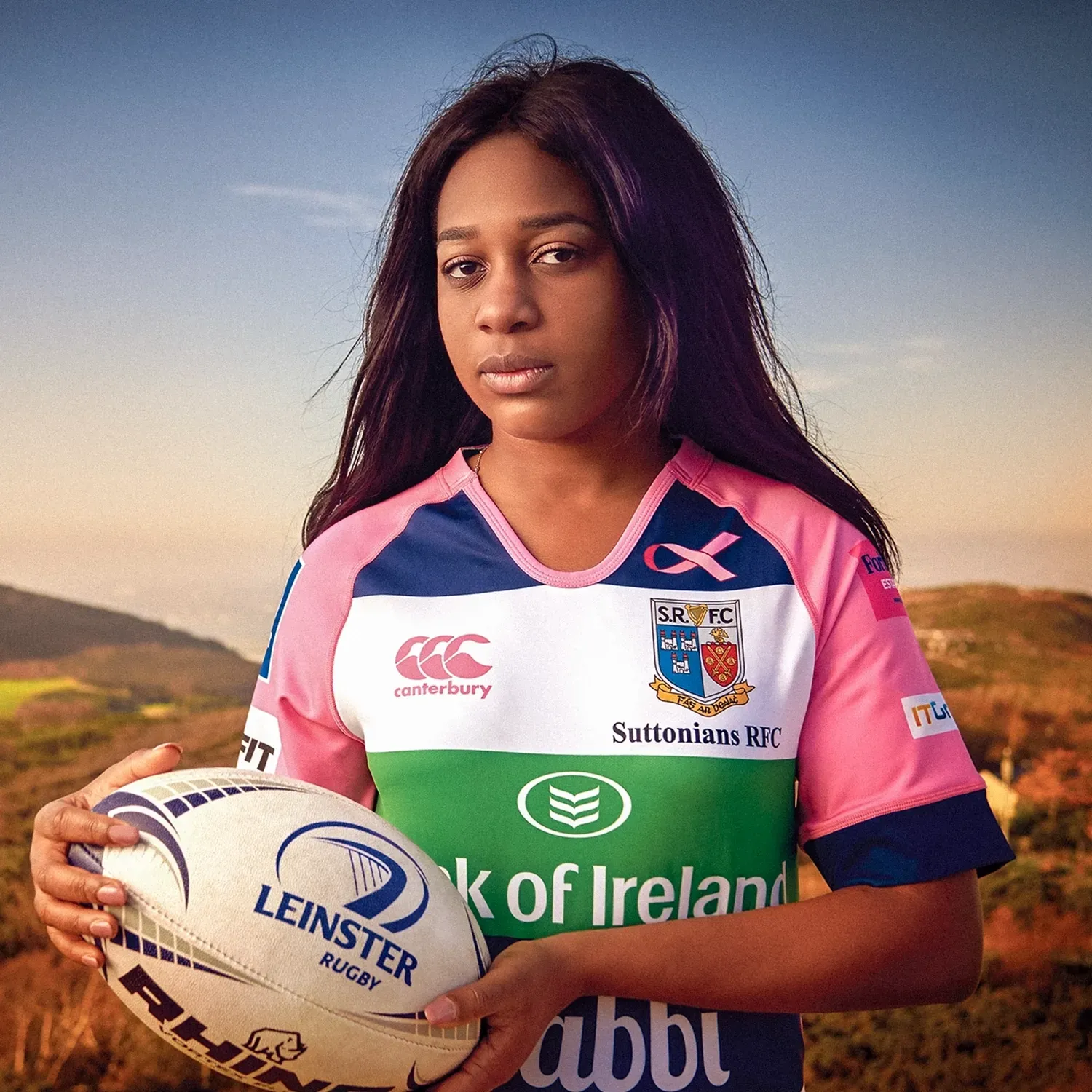WomanSuttonians rugby jersey holds a rugby ball, standing outdoors with a mountain backdrop.