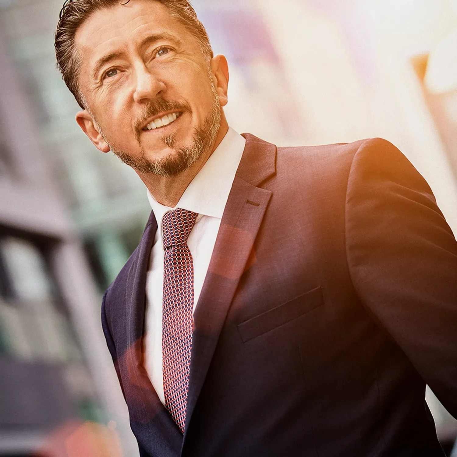 Smiling man in a suit, looking upwards, outdoors near a tall building.