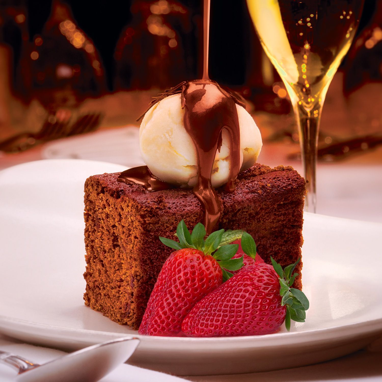 Chocolate cake with ice cream, chocolate sauce, and strawberries on a white plate with champagne in the background.