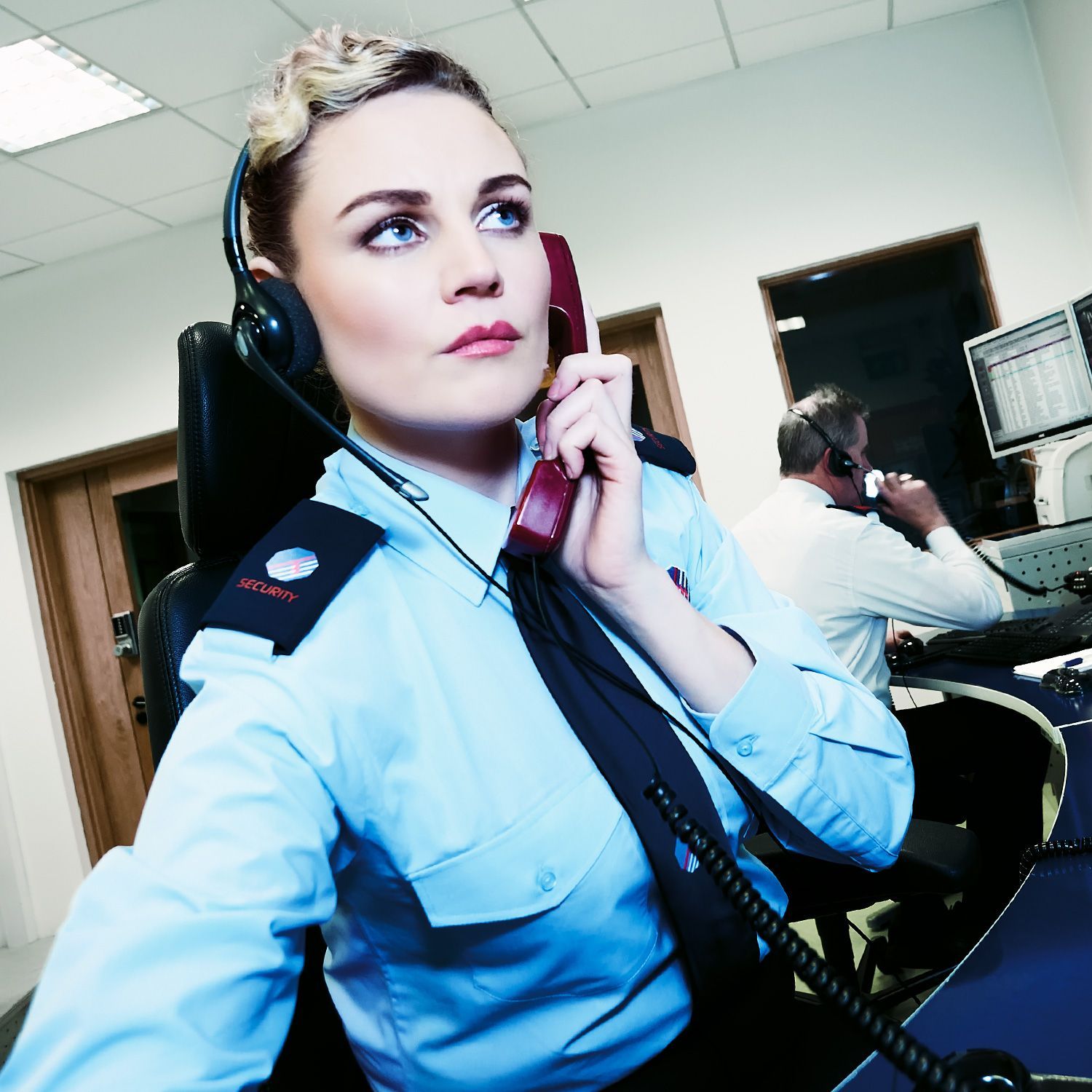 Woman in uniform with headset and phone, looking up thoughtfully in a control room.