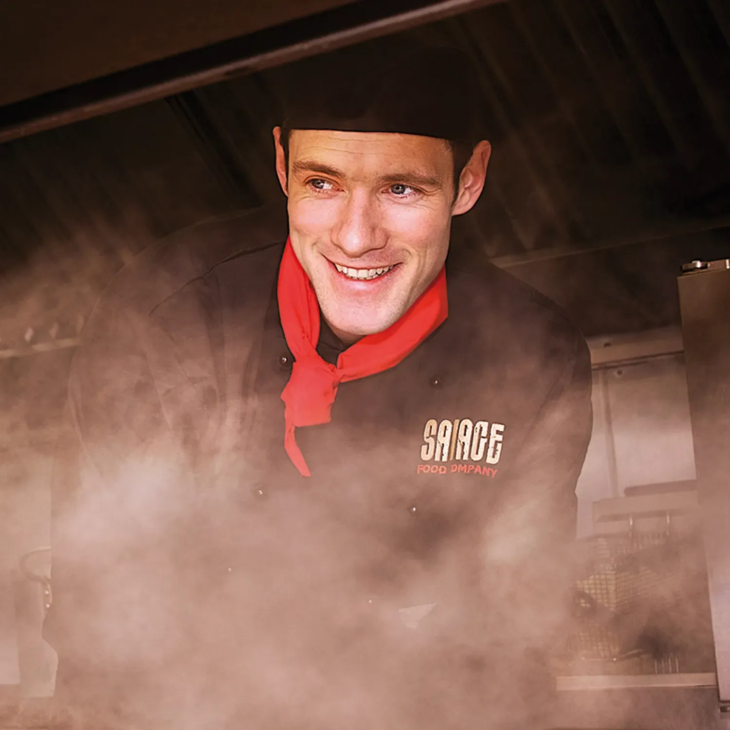 Chef smiling in kitchen, wearing black chef's coat, red neckerchief, and black cap, steam rising around him.