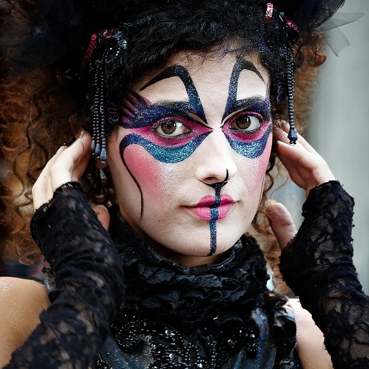 Woman with elaborate theatrical makeup and dark curly hair, hands on temples.