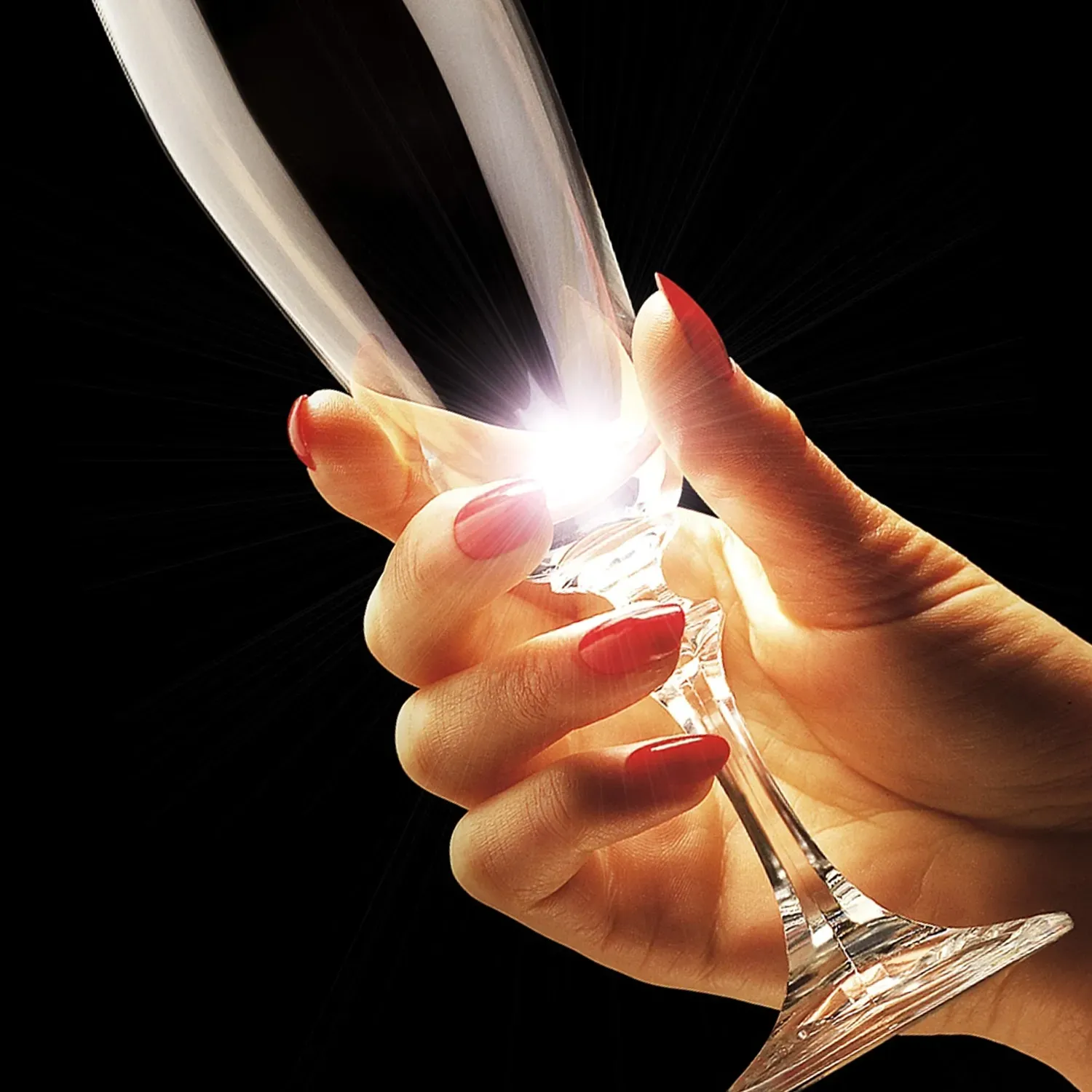 Hand with red nails holding a champagne flute against a black background, light shines through.