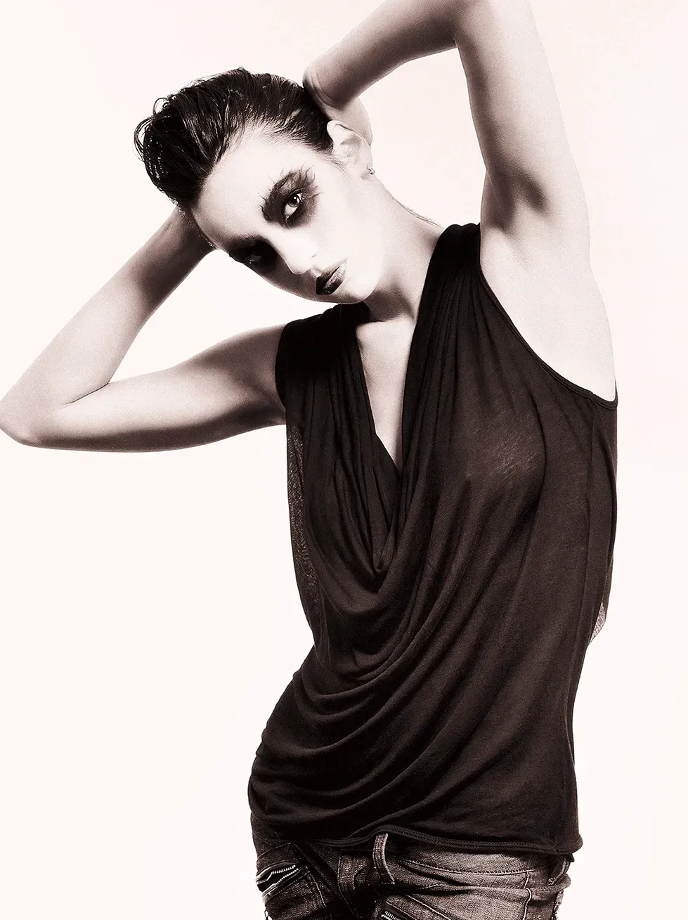 Woman with dark eye makeup, hands behind head, wearing a black top and jeans, against a white background.