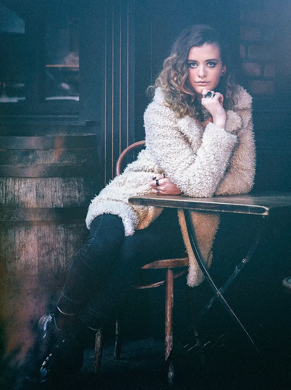 Woman seated at a cafe table, leaning on her arm, wearing a white coat.