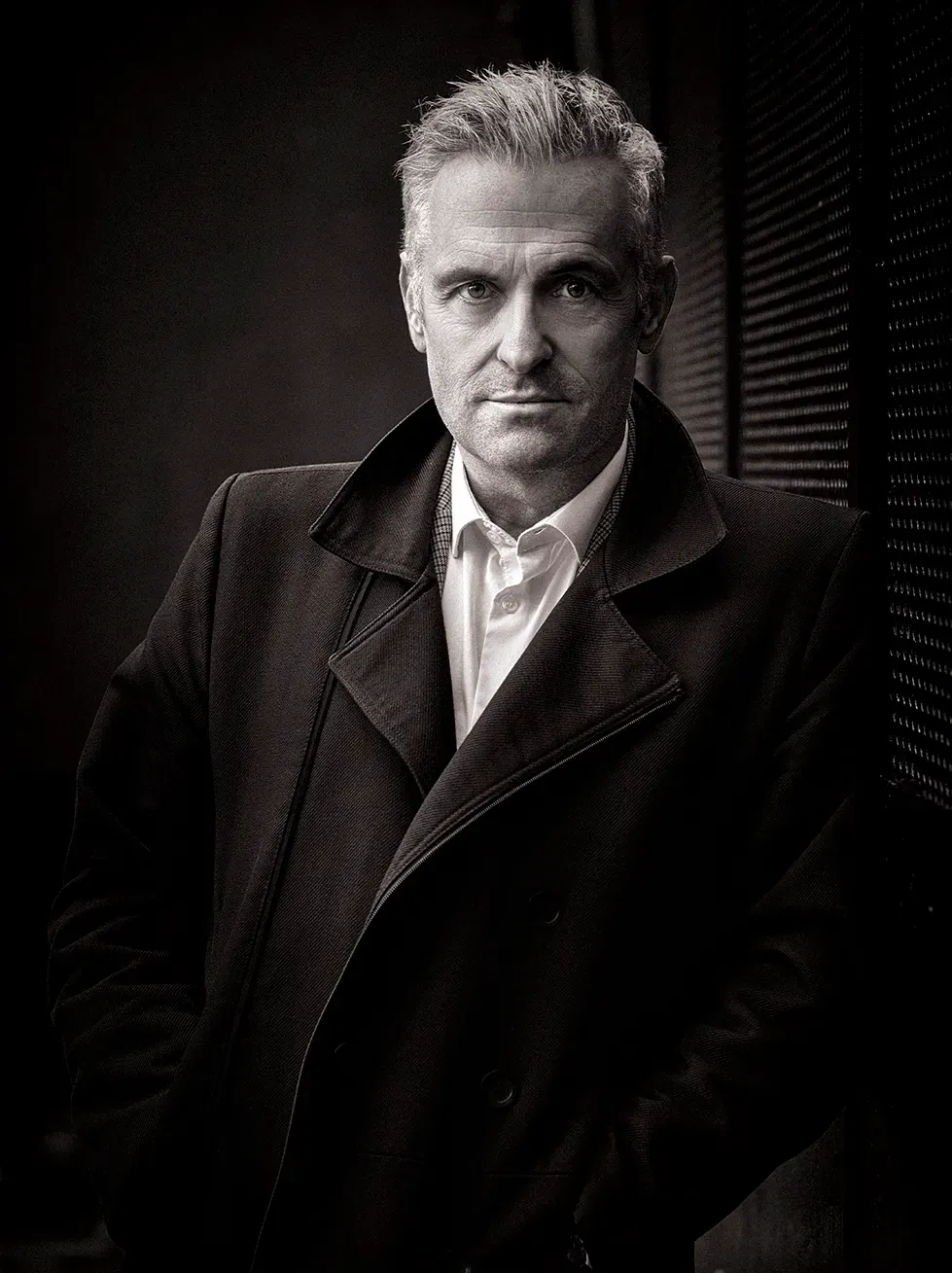 Man in dark coat, hands in pockets, looking directly at the viewer, dark background.